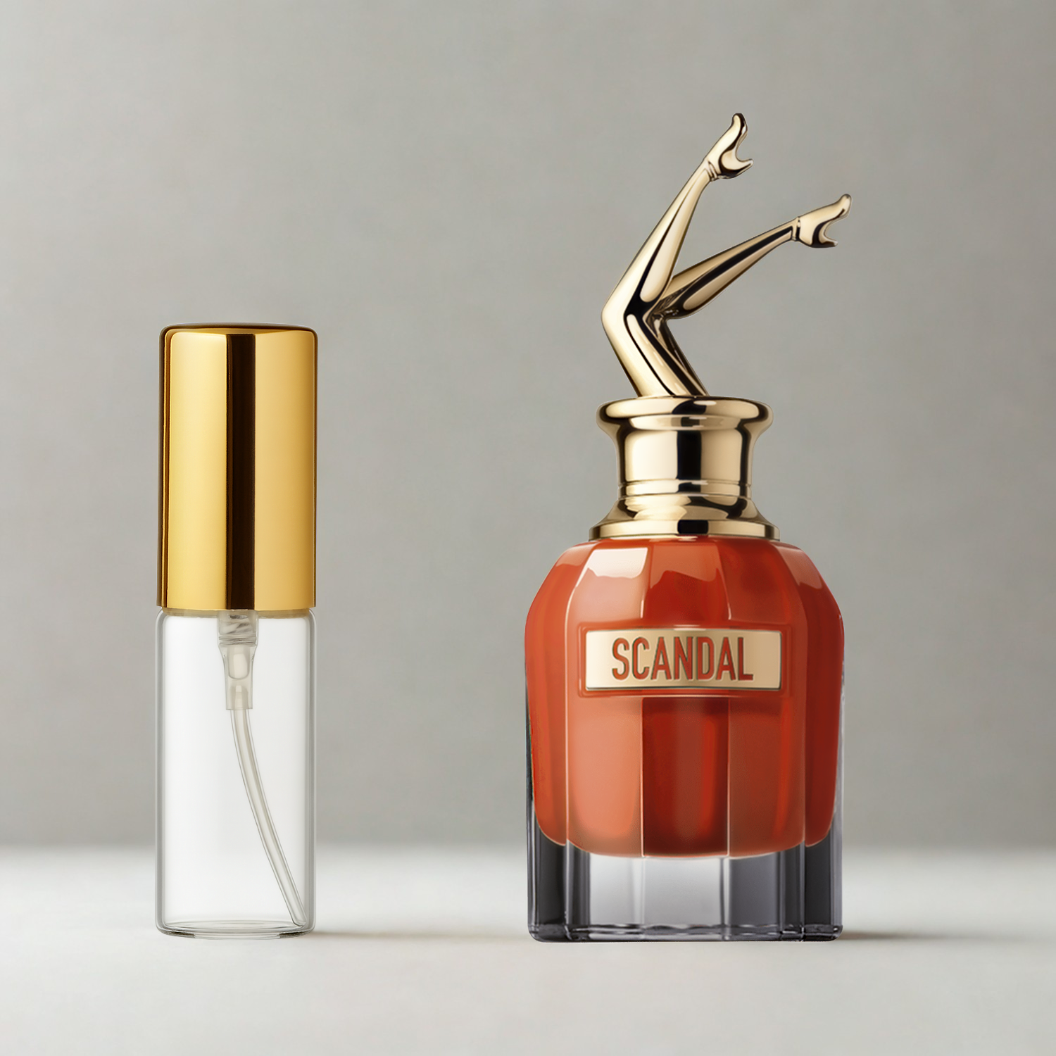 Decant de 5ml com borrifador dourado ao lado do frasco original do perfume importado feminino Scandal Le Parfum Jean Paul Gaultier Eau de Parfum, ambos sobre uma superfície e fundo cinza claro. A imagem destaca a apresentação do decant para venda e a familiaridade com o frasco original, disponíveis na Velyre.