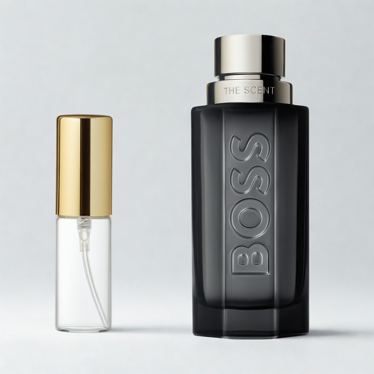 Decant de 5ml com borrifador dourado ao lado do frasco original do perfume importado masculino The Scent Magnetic Hugo Boss Eau de Parfum, ambos sobre uma superfície e fundo cinza claro. A imagem destaca a apresentação do decant para venda e a familiaridade com o frasco original, disponíveis na Velyre.