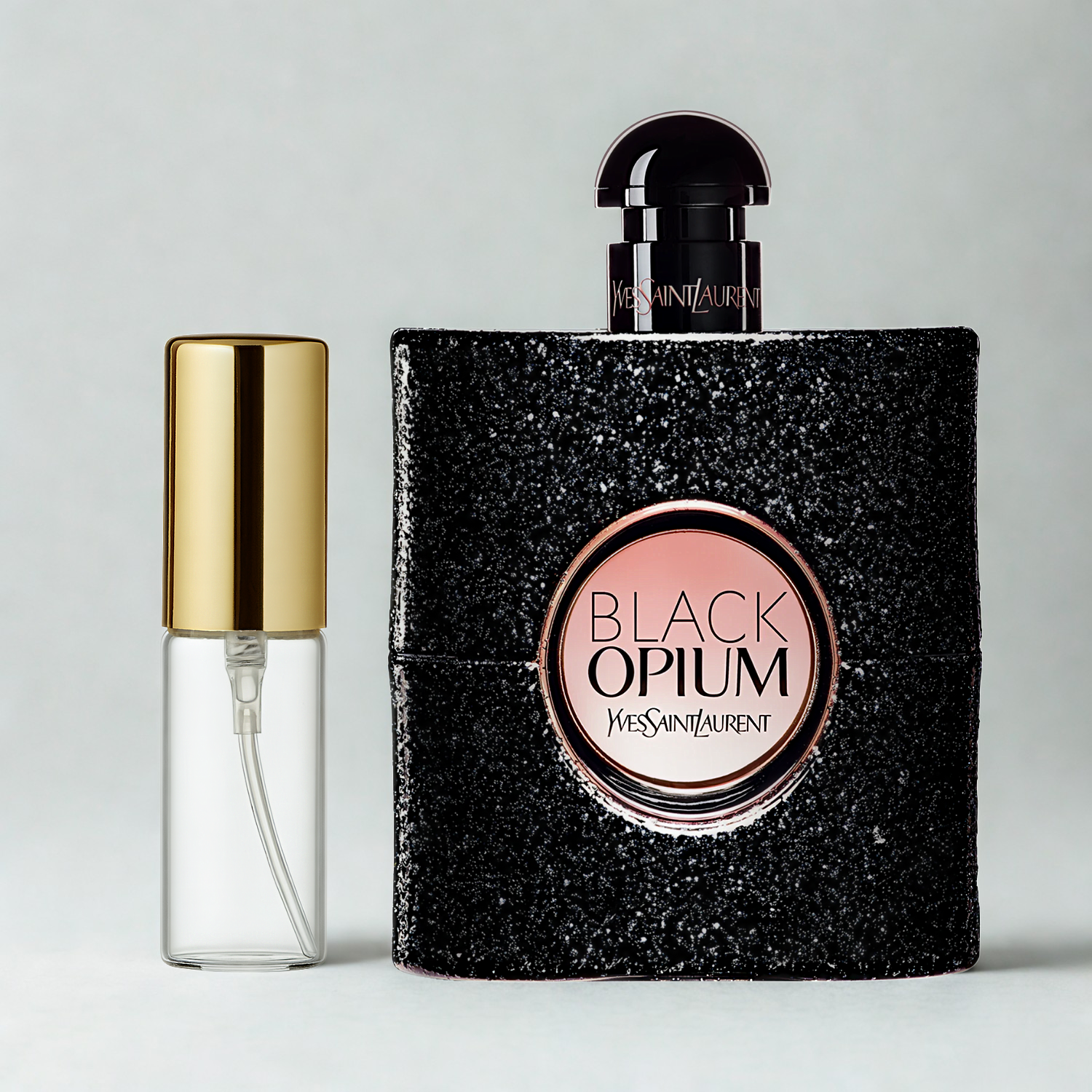 Decant de 5ml com borrifador dourado ao lado do frasco original do perfume importado feminino Black Opium Saint Laurent Eau de Parfum, ambos sobre uma superfície e fundo cinza claro. A imagem destaca a apresentação do decant para venda e a familiaridade com o frasco original, disponíveis na Velyre.