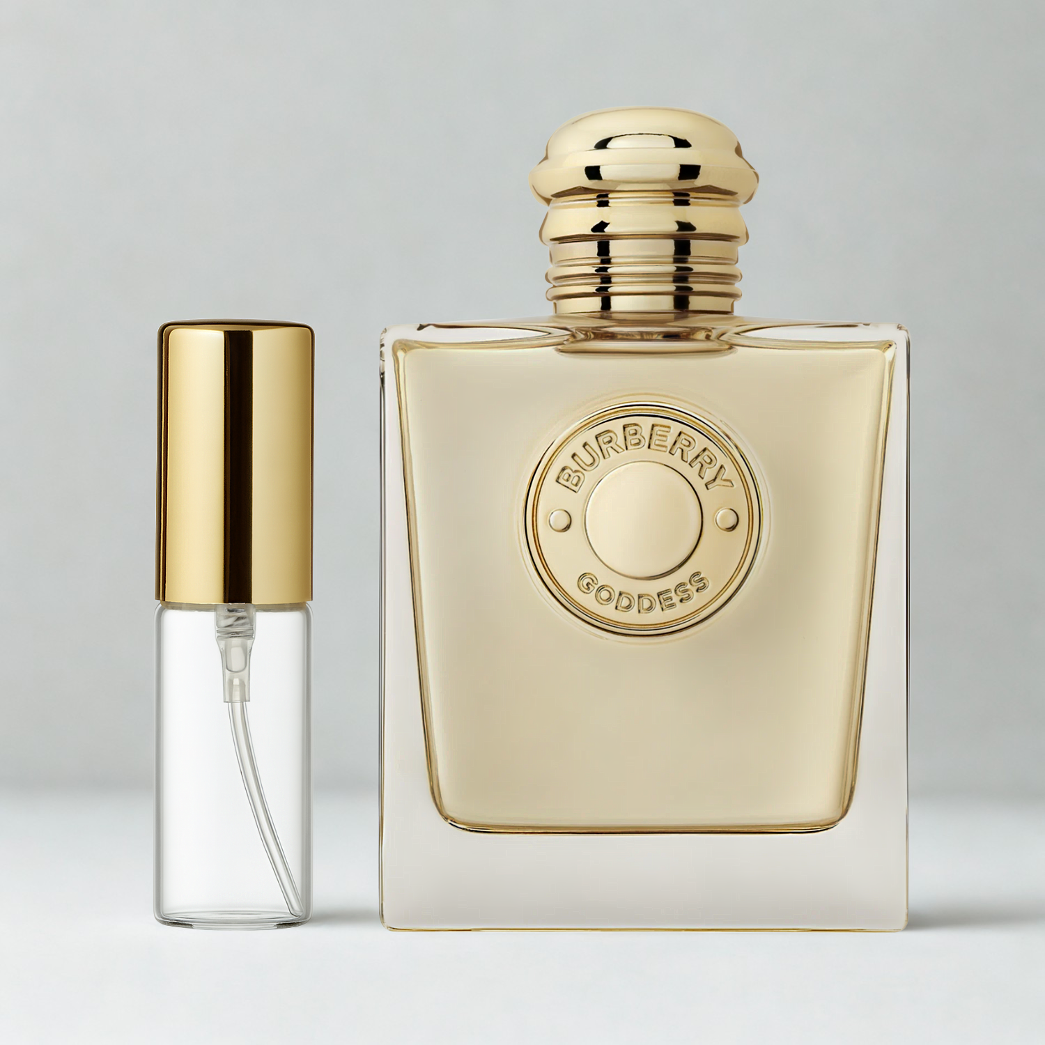 Decant de 5ml com borrifador dourado ao lado do frasco original do perfume importado feminino Goddess Burberry Eau de Parfum, ambos sobre uma superfície e fundo cinza claro. A imagem destaca a apresentação do decant para venda e a familiaridade com o frasco original, disponíveis na Velyre.