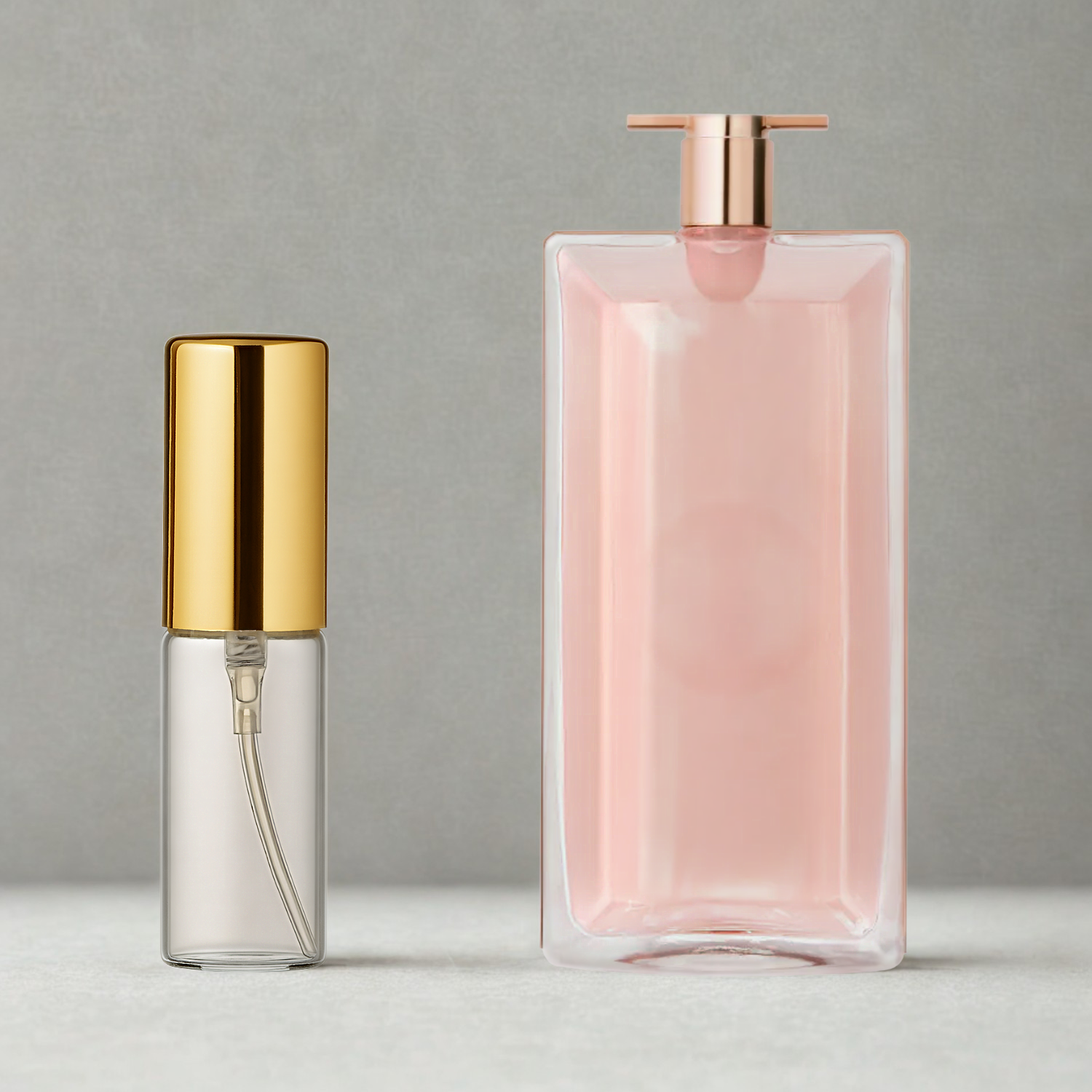 Decant de 5ml com borrifador dourado ao lado do frasco original do perfume importado feminino Idôle Lancôme Eau de Parfum, ambos sobre uma superfície e fundo cinza claro. A imagem destaca a apresentação do decant para venda e a familiaridade com o frasco original, disponíveis na Velyre.