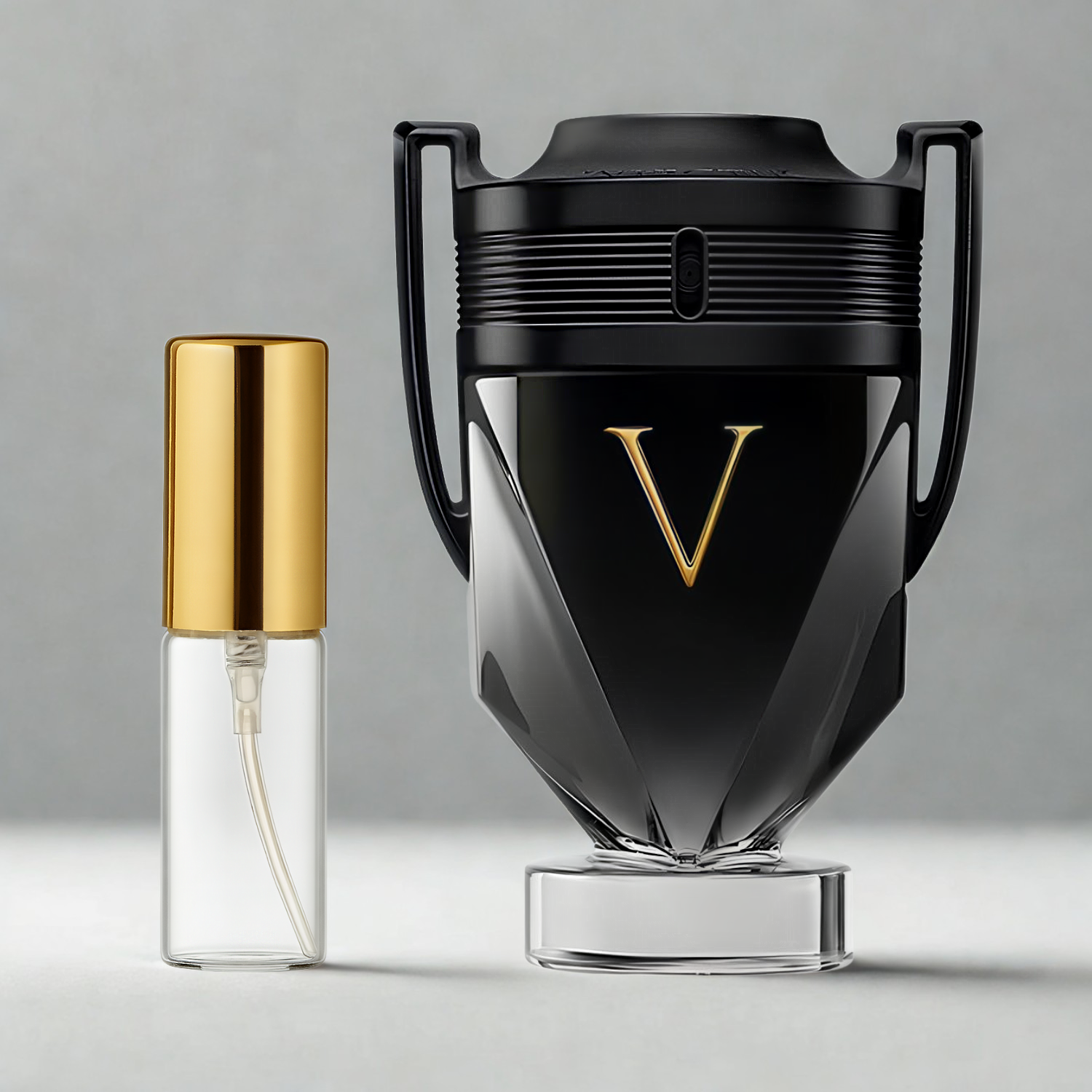 Decant de 5ml com borrifador dourado ao lado do frasco original do perfume importado masculino Invictus Victory Rabanne Eau de Parfum, ambos sobre uma superfície e fundo cinza claro. A imagem destaca a apresentação do decant para venda e a familiaridade com o frasco original, disponíveis na Velyre.