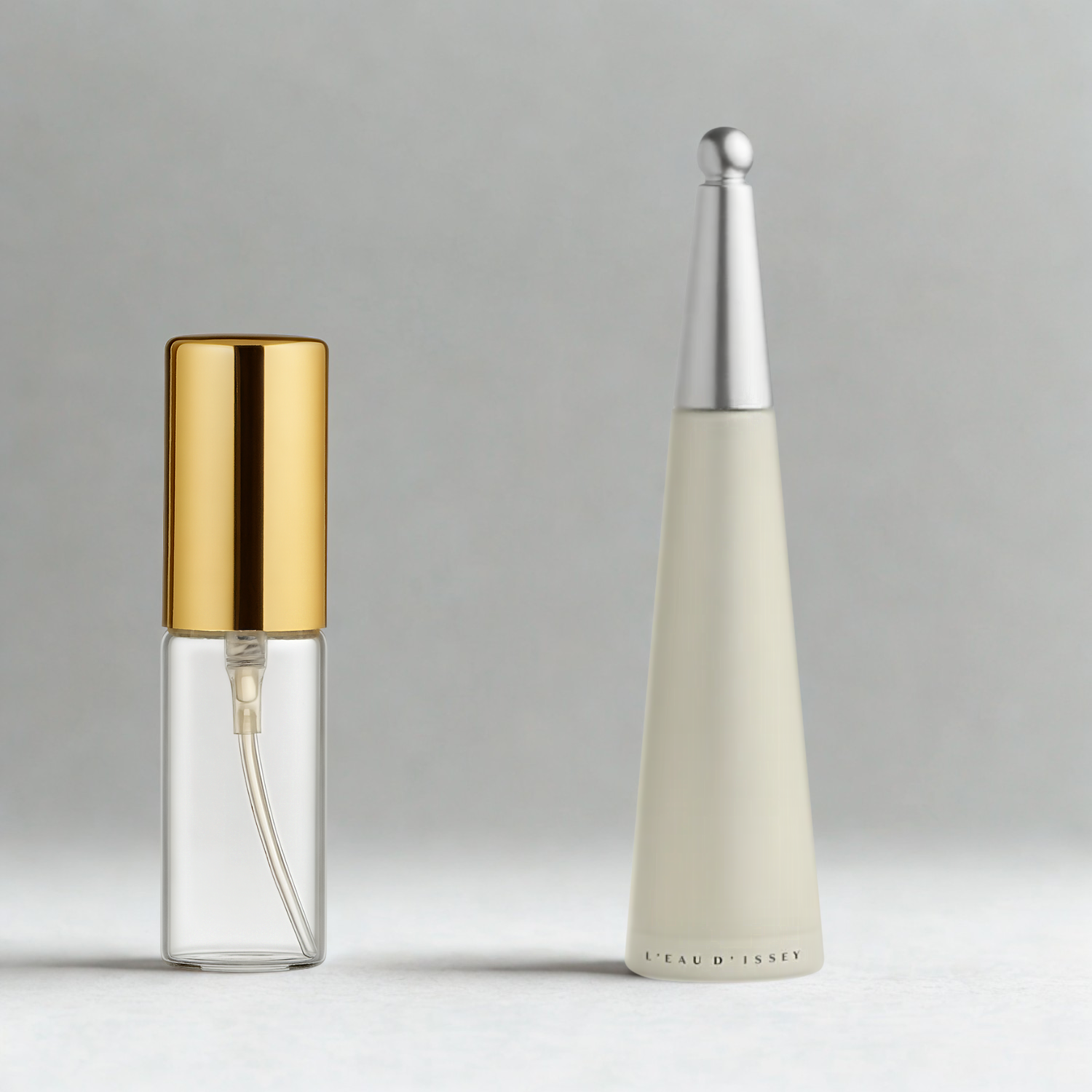 Decant de 5ml com borrifador dourado ao lado do frasco original do perfume importado feminino L'Eau d'Issey Issey Miyake Eau de Toilette, ambos sobre uma superfície e fundo cinza claro. A imagem destaca a apresentação do decant para venda e a familiaridade com o frasco original, disponíveis na Velyre.