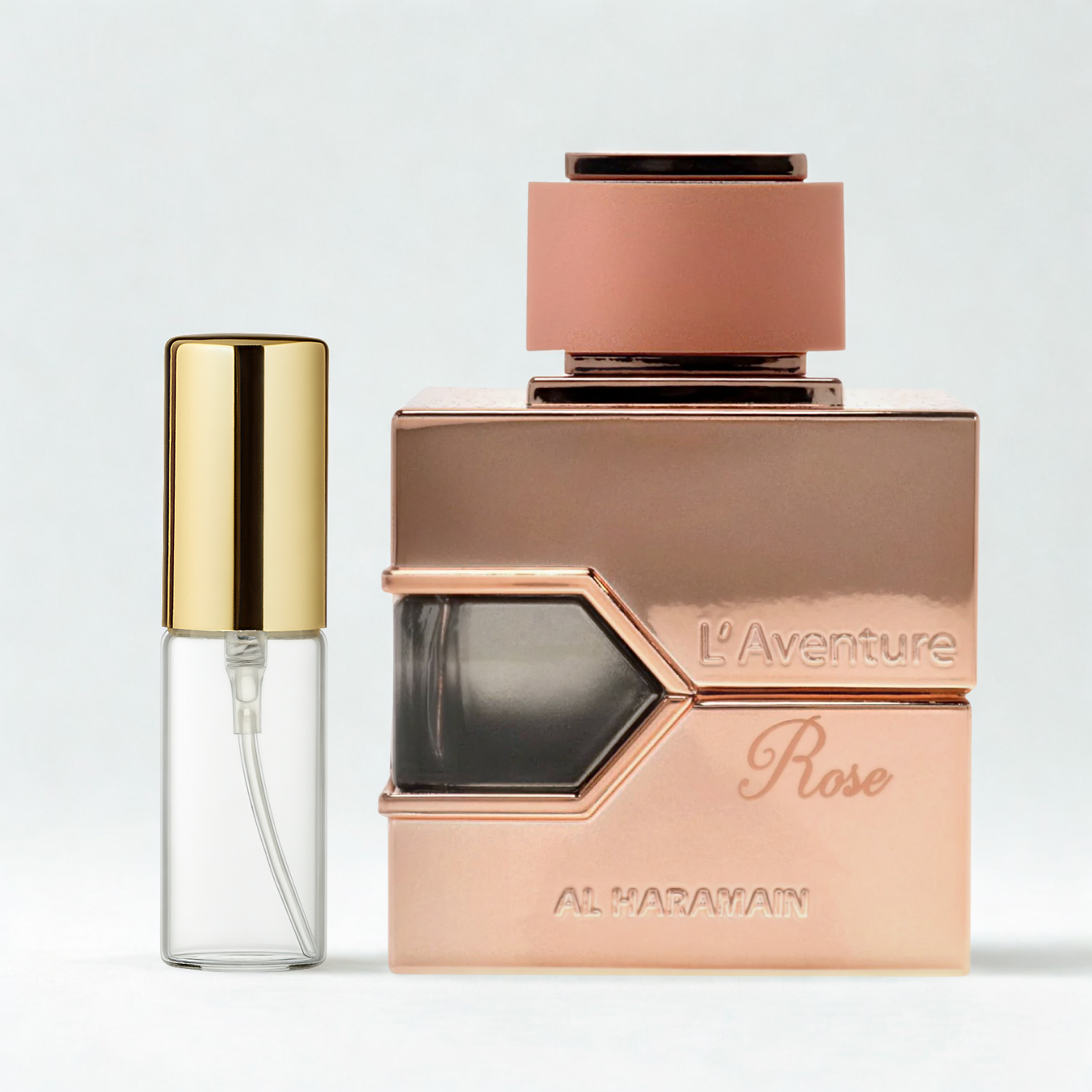 Decant de 5ml com borrifador dourado ao lado do frasco original do perfume árabe feminino L'aventure Rose Al Haramain Eau de Parfum, ambos sobre uma superfície e fundo cinza claro. A imagem destaca a apresentação do decant para venda e a familiaridade com o frasco original, disponíveis na Velyre.