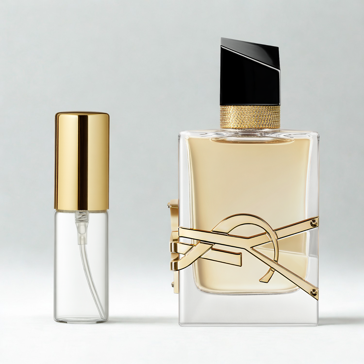 Decant de 5ml com borrifador dourado ao lado do frasco original do perfume importado feminino Libre Saint Laurent Eau de Parfum, ambos sobre uma superfície e fundo cinza claro. A imagem destaca a apresentação do decant para venda e a familiaridade com o frasco original, disponíveis na Velyre.