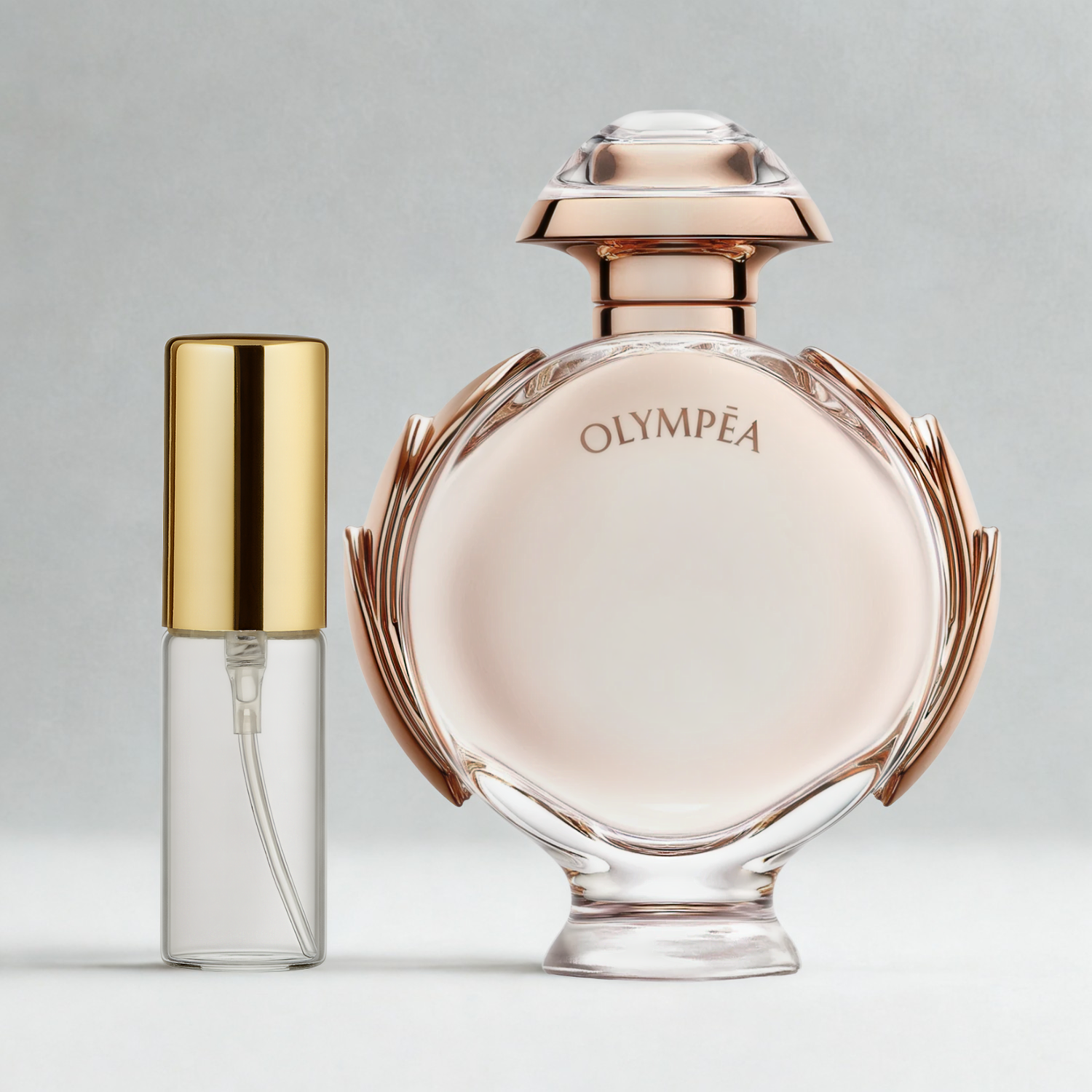 Decant de 5ml com borrifador dourado ao lado do frasco original do perfume importado feminino Olympéa Rabanne Eau de Parfum, ambos sobre uma superfície e fundo cinza claro. A imagem destaca a apresentação do decant para venda e a familiaridade com o frasco original, disponíveis na Velyre.