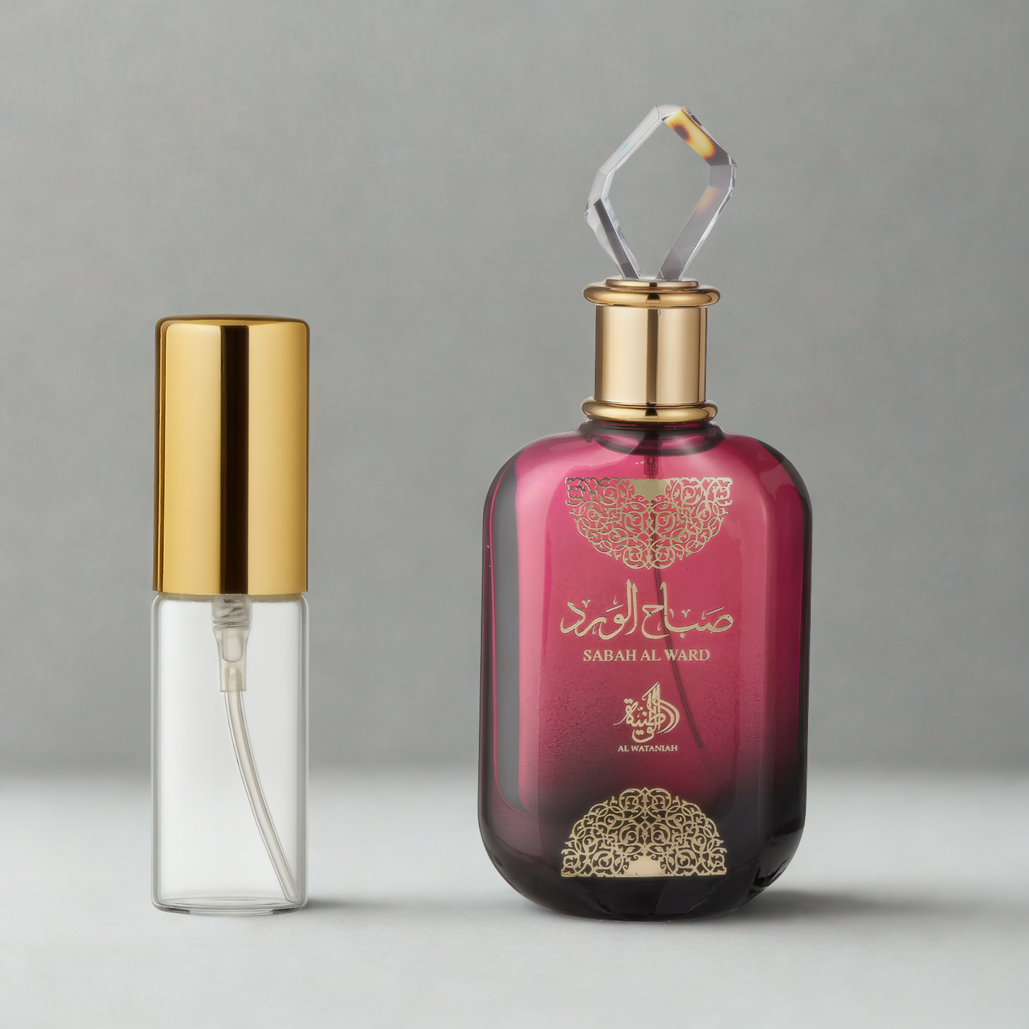 Decant de 5ml com borrifador dourado ao lado do frasco original do perfume árabe feminino Sabah Al Ward Al Wataniah Eau de Parfum, ambos sobre uma superfície e fundo cinza claro. A imagem destaca a apresentação do decant para venda e a familiaridade com o frasco original, disponíveis na Velyre.