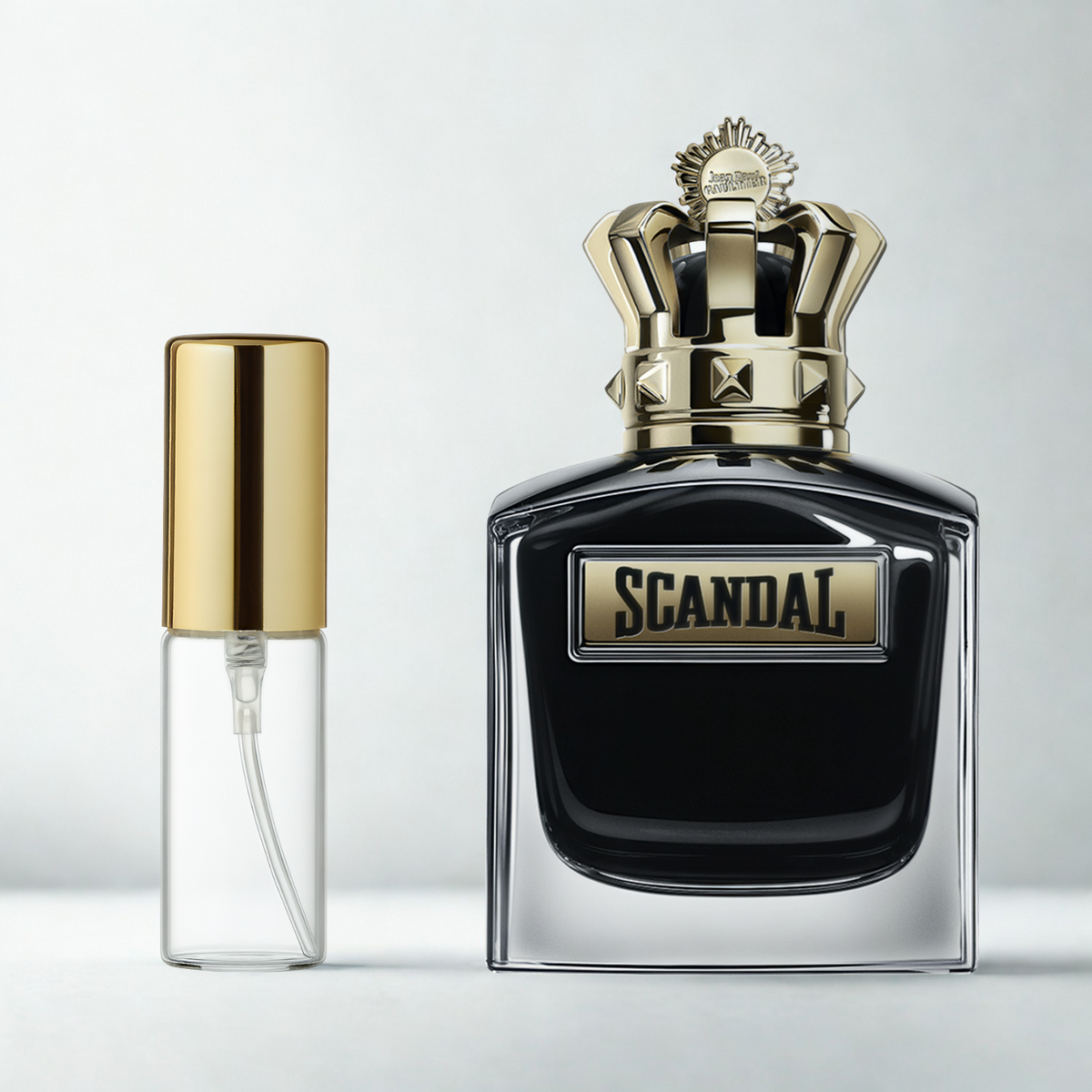 Decant de 5ml com borrifador dourado ao lado do frasco original do perfume importado masculino Scandal Pour Homme Le Parfum Jean Paul Gaultier Eau de Parfum, ambos sobre uma superfície e fundo cinza claro. A imagem destaca a apresentação do decant para venda e a familiaridade com o frasco original, disponíveis na Velyre.