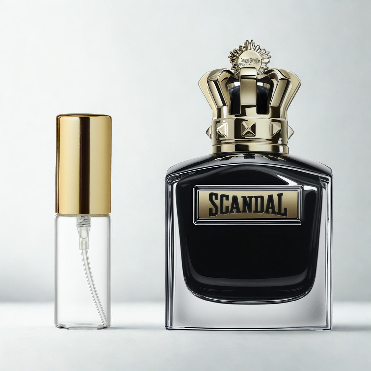 Decant de 5ml com borrifador dourado ao lado do frasco original do perfume importado masculino Scandal Pour Homme Le Parfum Jean Paul Gaultier Eau de Parfum, ambos sobre uma superfície e fundo cinza claro. A imagem destaca a apresentação do decant para venda e a familiaridade com o frasco original, disponíveis na Velyre.