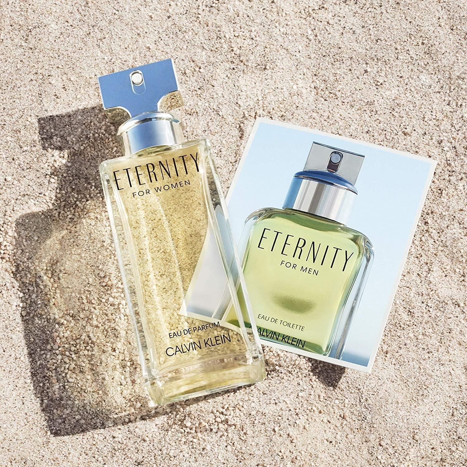 Dois frascos dos perfumes Eternity Calvin Klein, a versão feminina com líquido amarelo pálido e a masculina com líquido esverdeado, lado a lado sobre uma superfície arenosa com uma luz que projeta suas sombras, sugerindo um casal unido pela eternidade.