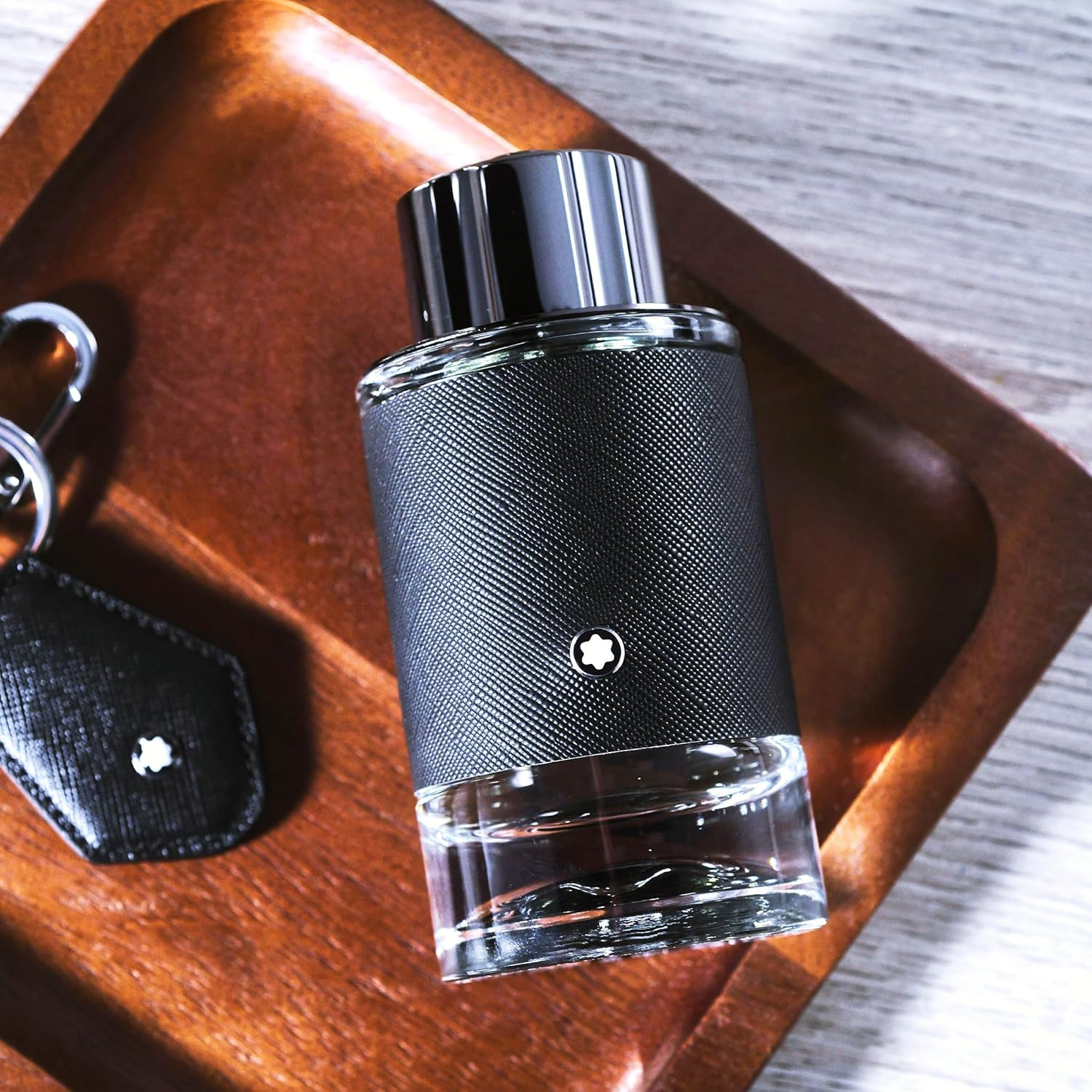 Perfume Explorer Montblanc posicionado em uma bandeja de madeira escura ao lado de um chaveiro de couro com o logo da marca, transmitindo uma sensação de luxo discreto e viagem.
