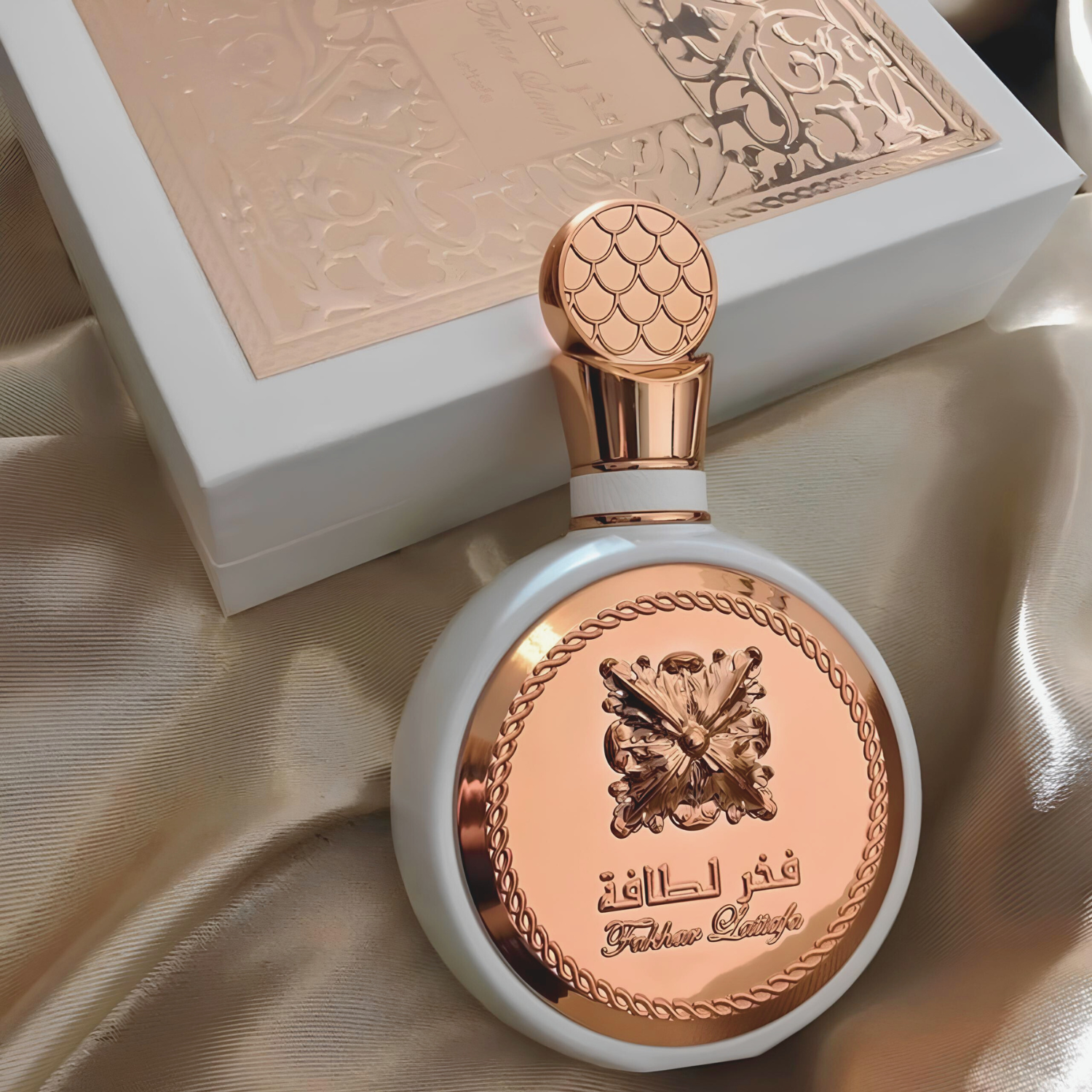 Perfume árabe Fakhar Rose Lattafa, disponível na Velyre, posicionado sobre tecido acetinado dourado, com a caixa do perfume ao fundo, realçando a embalagem luxuosa e a sofisticação da fragrância.