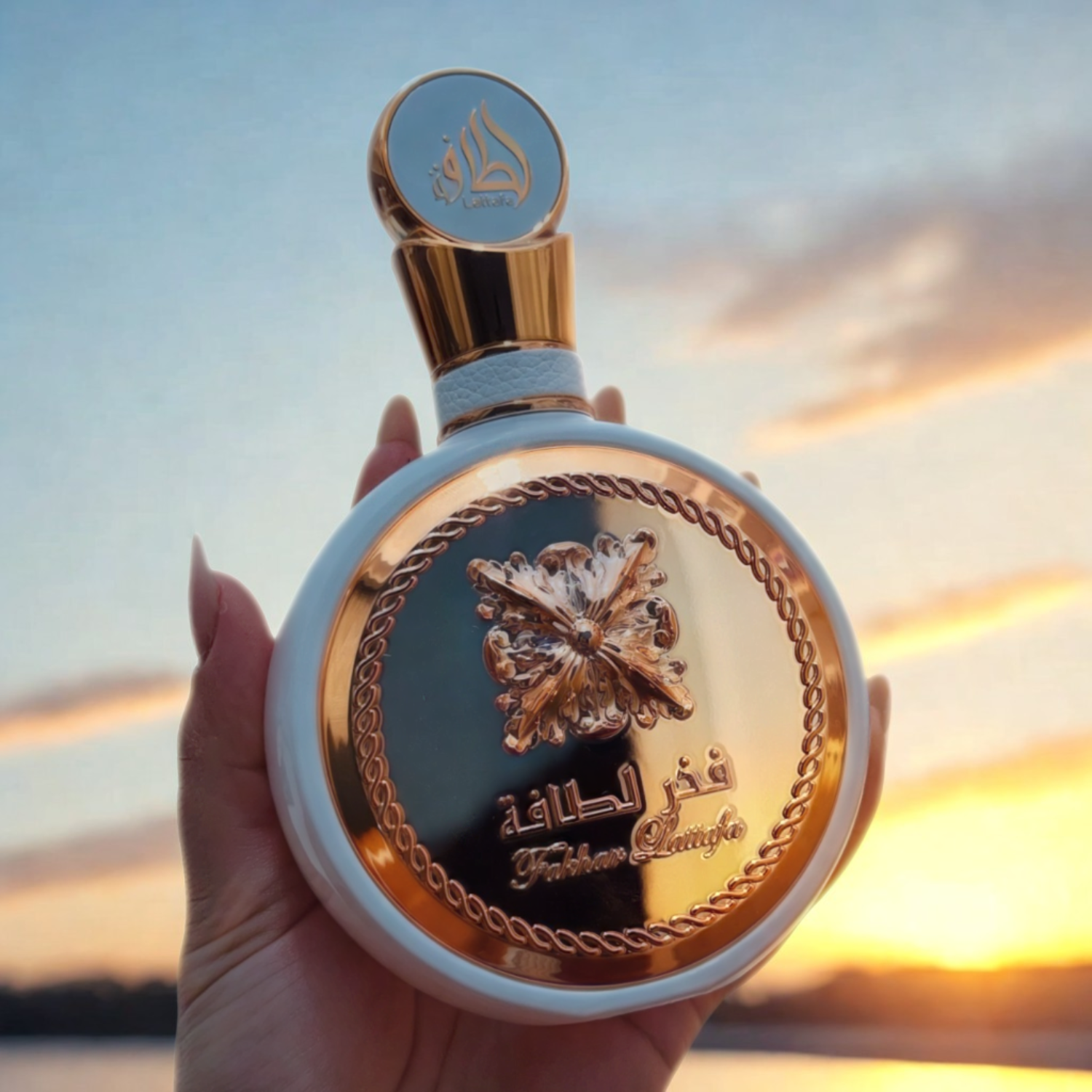 Mão feminina segurando o perfume árabe Fakhar Rose Lattafa, disponível na Velyre, contra um pôr do sol alaranjado e um céu com nuvens, evocando beleza e serenidade.