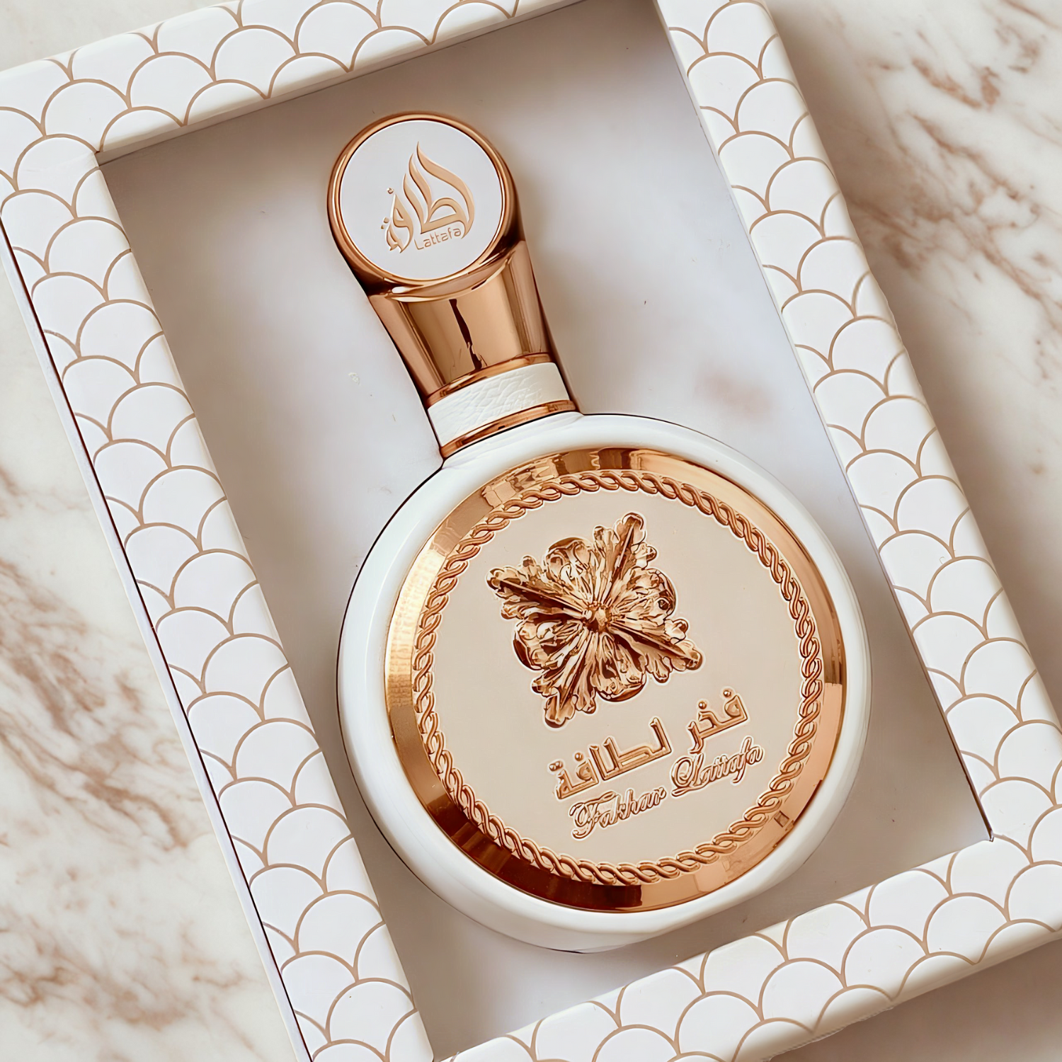 Perfume árabe Fakhar Rose Lattafa, disponível na Velyre, dentro de uma caixa branca com moldura decorativa em escala de peixe, destacando a apresentação elegante e os detalhes dourados do frasco.