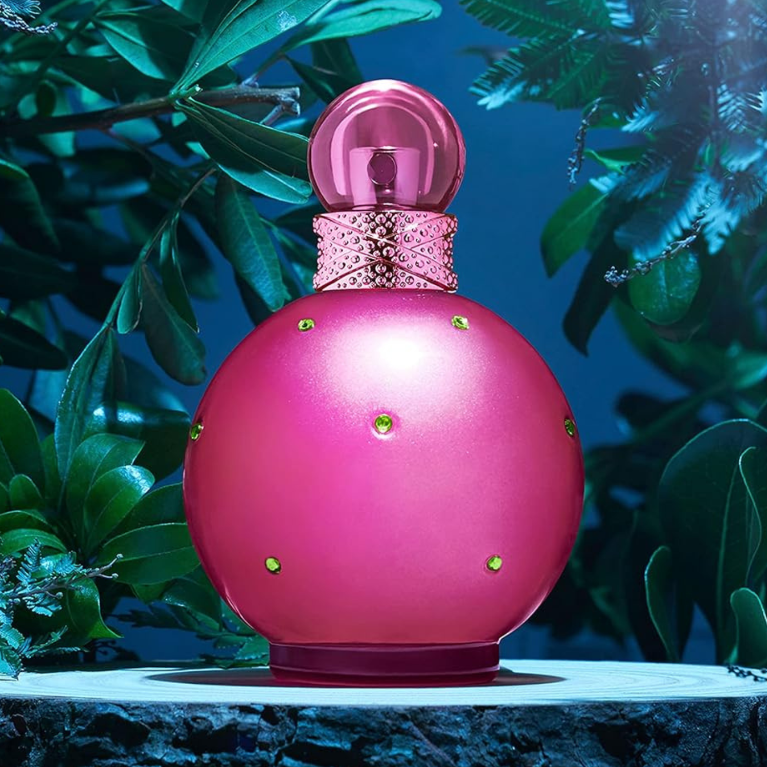 Frasco do perfume Fantasy de Britney Spears em destaque sobre uma base de madeira, cercado por folhas verdes. O frasco é rosa vibrante, em formato esférico, com detalhes em pedras verdes e tampa arredondada. Imagem com fundo azul escuro e iluminação suave.