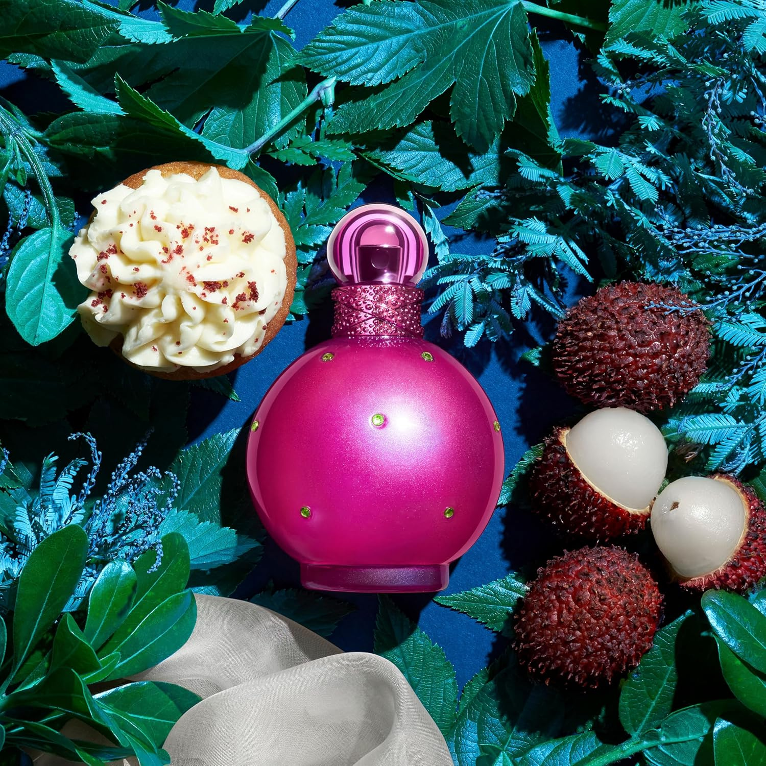 Perfume Fantasy de Britney Spears sobre fundo azul com folhas verdes. O frasco rosa está cercado por frutas lichia e um cupcake com cobertura branca, representando notas gourmand da fragrância.