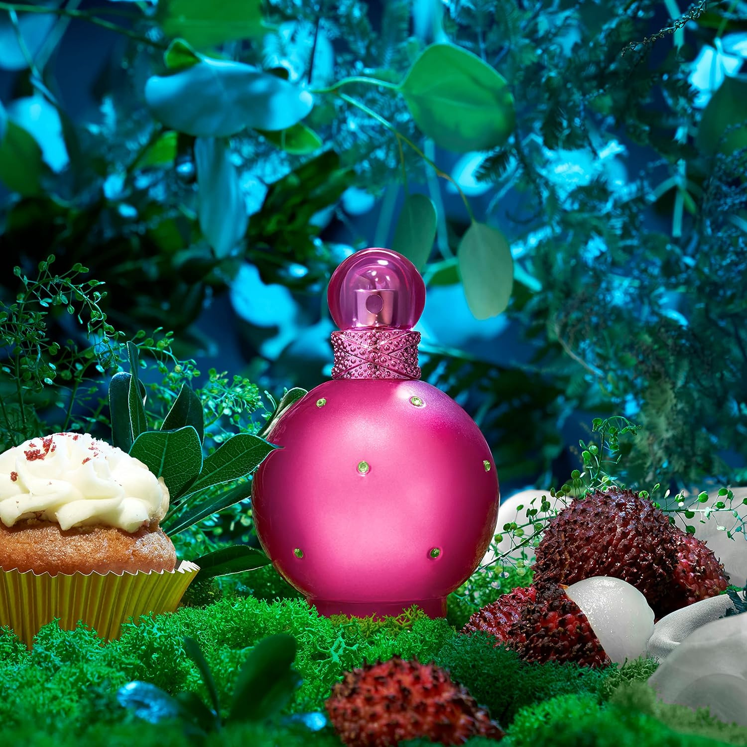 Perfume Fantasy de Britney Spears sobre musgo verde em meio a folhagens. O frasco rosa está acompanhado de lichias abertas e um cupcake com cobertura cremosa, em um cenário natural com iluminação suave.