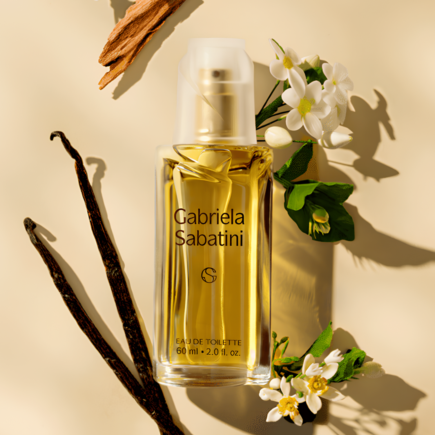 Perfume Gabriela Sabatini deitado sobre superfície bege, cercado por flores brancas, favas de baunilha e um pedaço de madeira. A composição destaca as notas olfativas da fragrância.