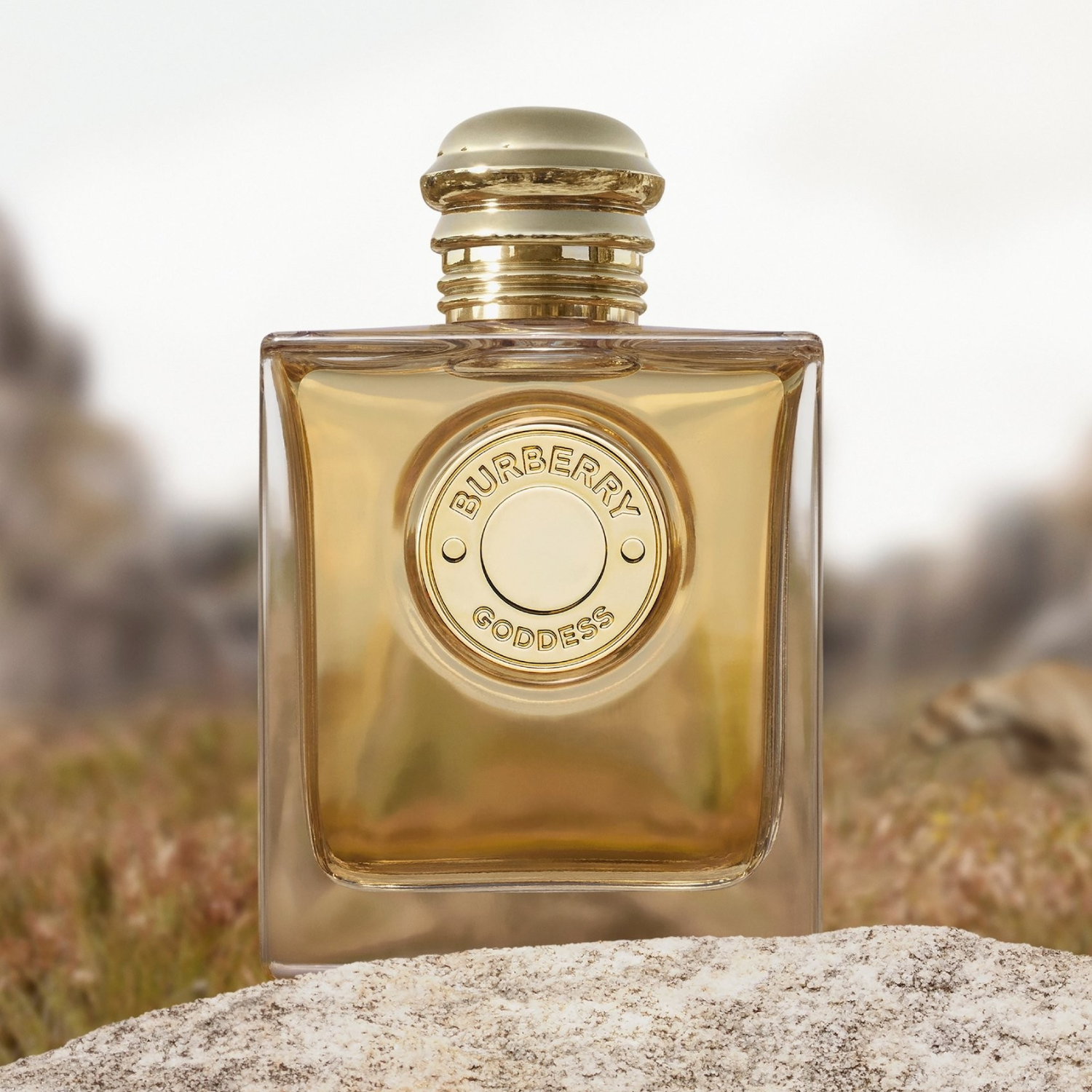 Perfume Burberry Goddess sobre uma pedra em ambiente natural, com fundo desfocado de montanhas. O frasco dourado reflete a luz suave do dia.