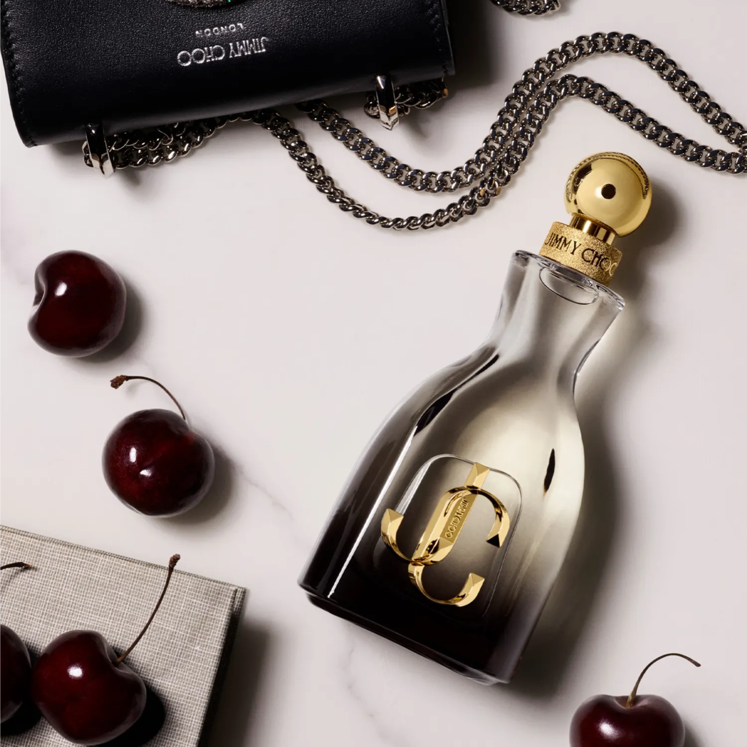 Perfume I Want Choo Forever Jimmy Choo em uma composição com uma pequena bolsa preta com corrente prata e cerejas vermelhas espalhadas sobre uma superfície de mármore branco, transmitindo uma atmosfera sofisticada e feminina com um toque de doçura.