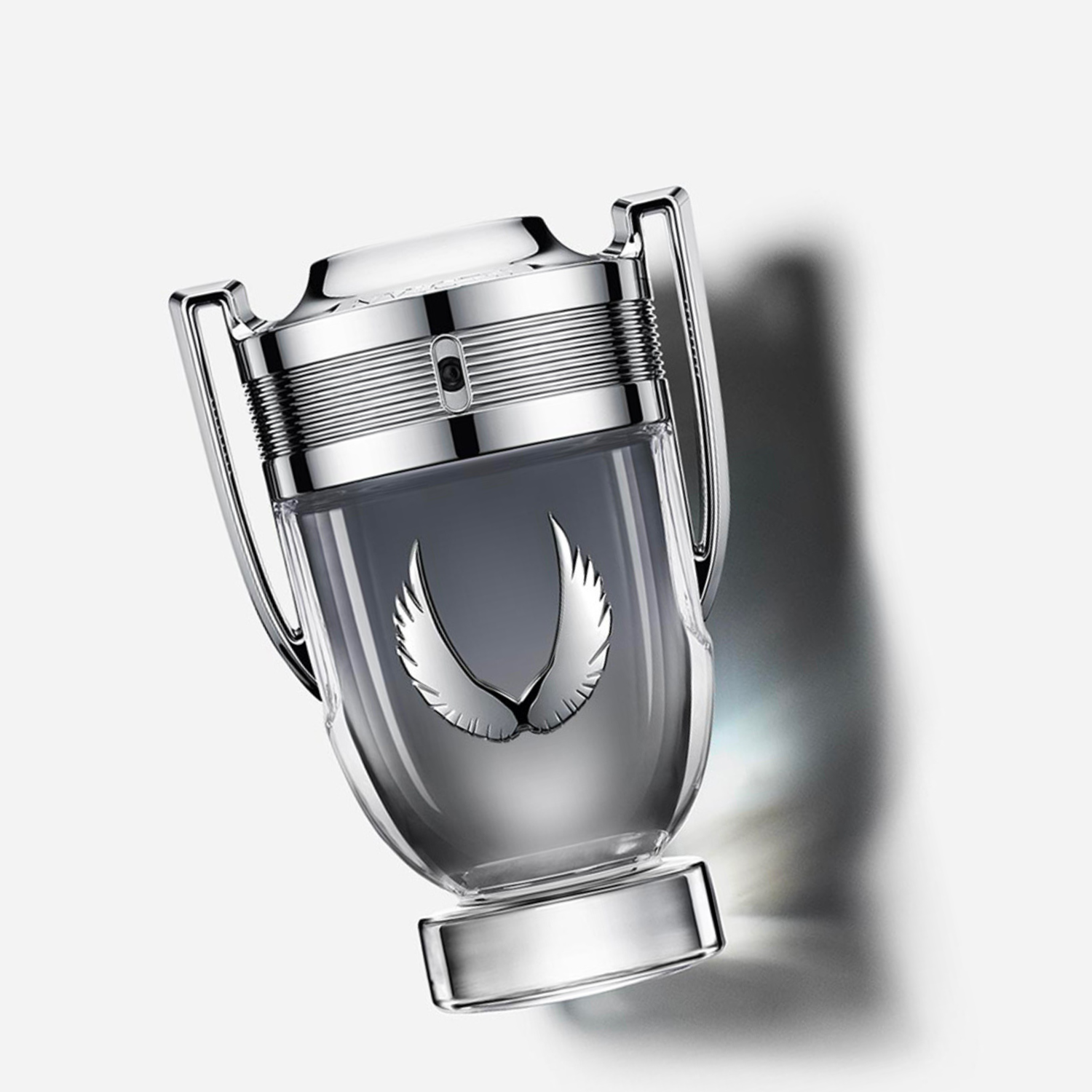 Perfume Invictus Platinum Rabanne inclinado sobre uma superfície clara com uma sombra dramática ao fundo, realçando o design do troféu e as asas prateadas, transmitindo uma sensação de vitória e elegância.