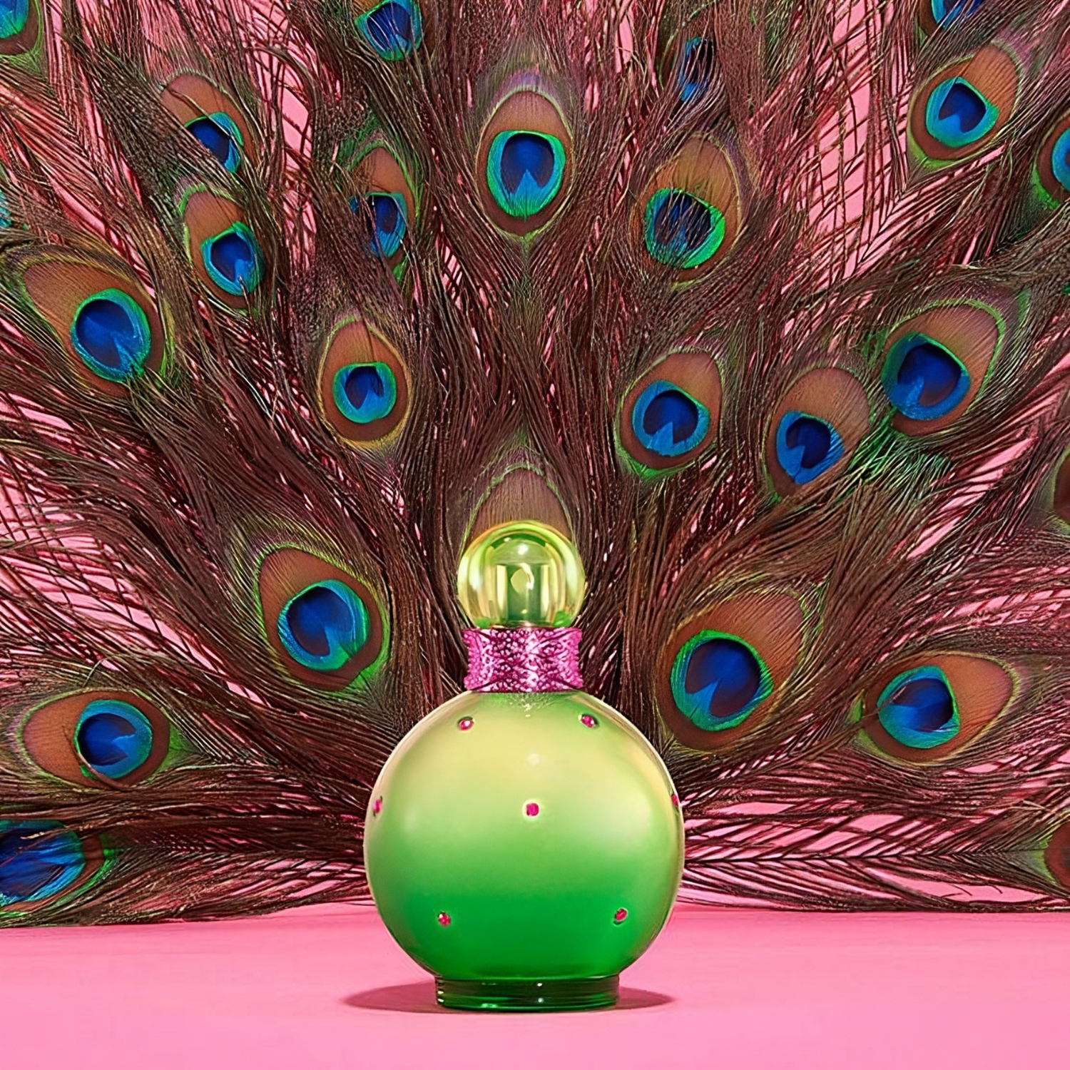 Perfume Jungle Fantasy de Britney Spears posicionado em frente a penas de pavão abertas sobre fundo rosa. O frasco verde contrasta com os tons intensos azul e verde das penas, criando uma composição tropical.