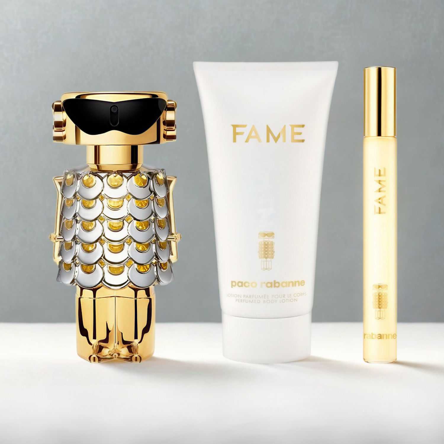 Kit feminino importado Fame Rabanne Eau de Parfum, disponível na Velyre, com perfume de 50ml em frasco de robô dourado, caneta de 10ml e hidratante de 75ml, dispostos lado a lado sobre uma superfície cinza clara.