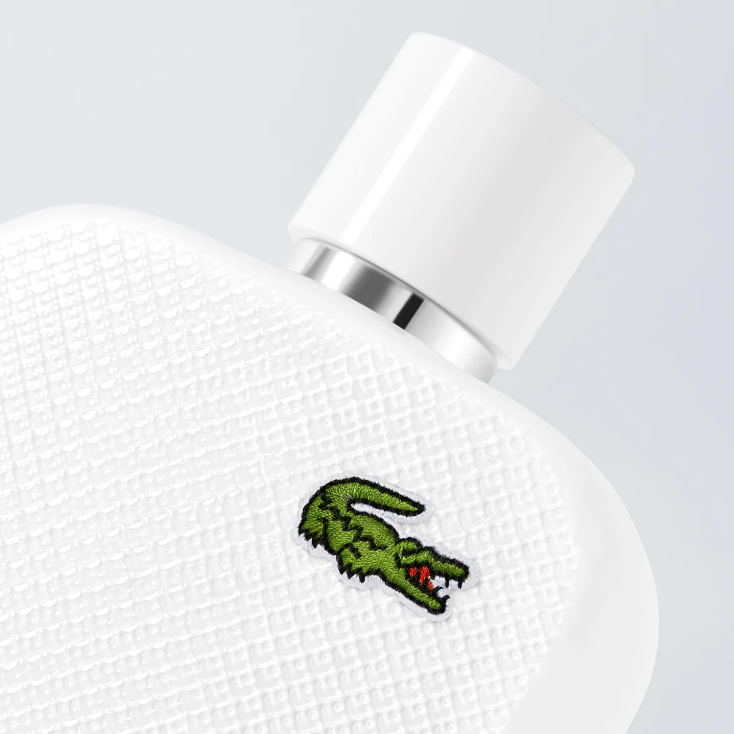Close-up do frasco branco texturizado do perfume L.12.12 Blanc Lacoste, destacando o icônico logo de crocodilo verde da marca.
