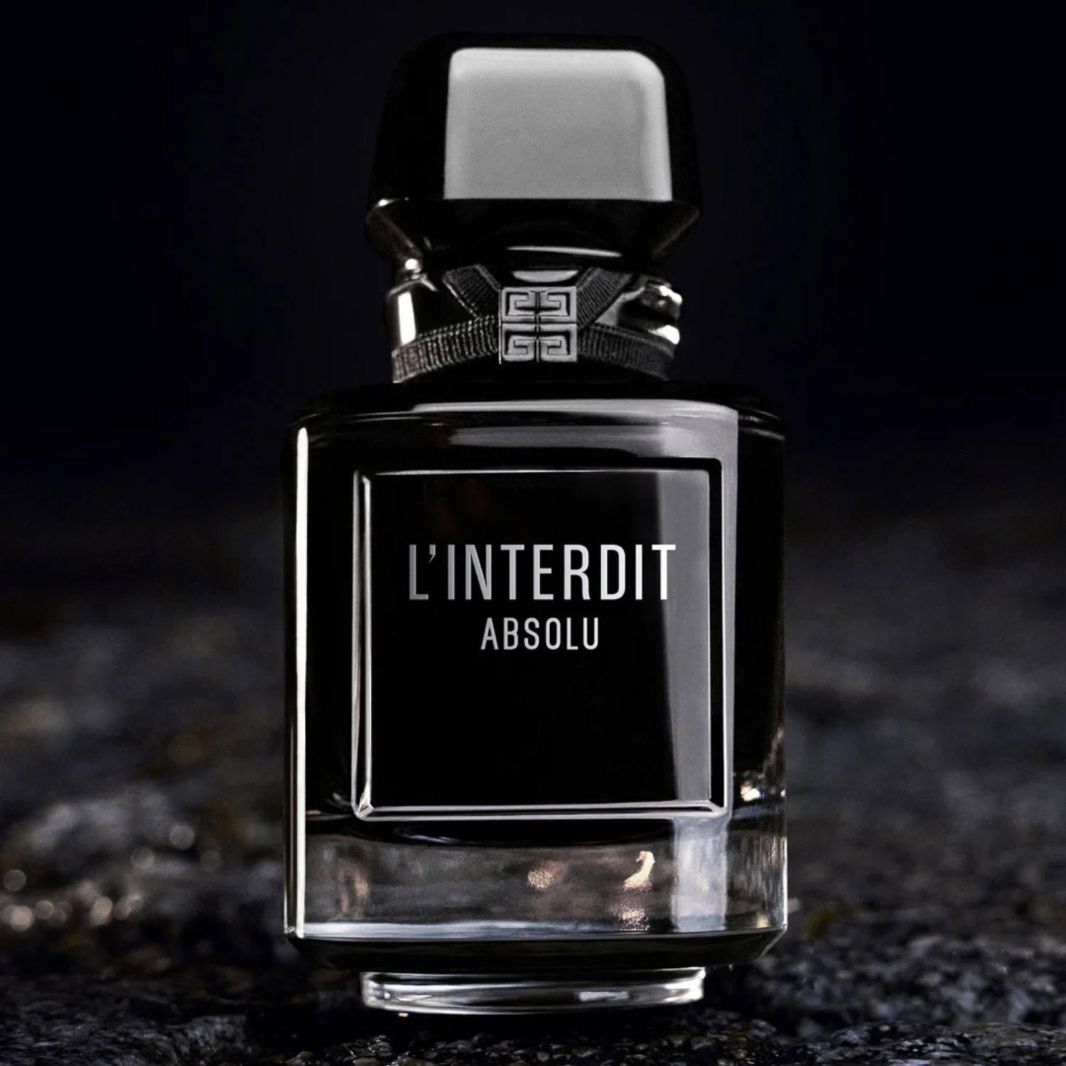 Close do perfume L’Interdit Absolu sobre superfície escura, com reflexos de luz que realçam o vidro preto e o contraste com a escrita branca.