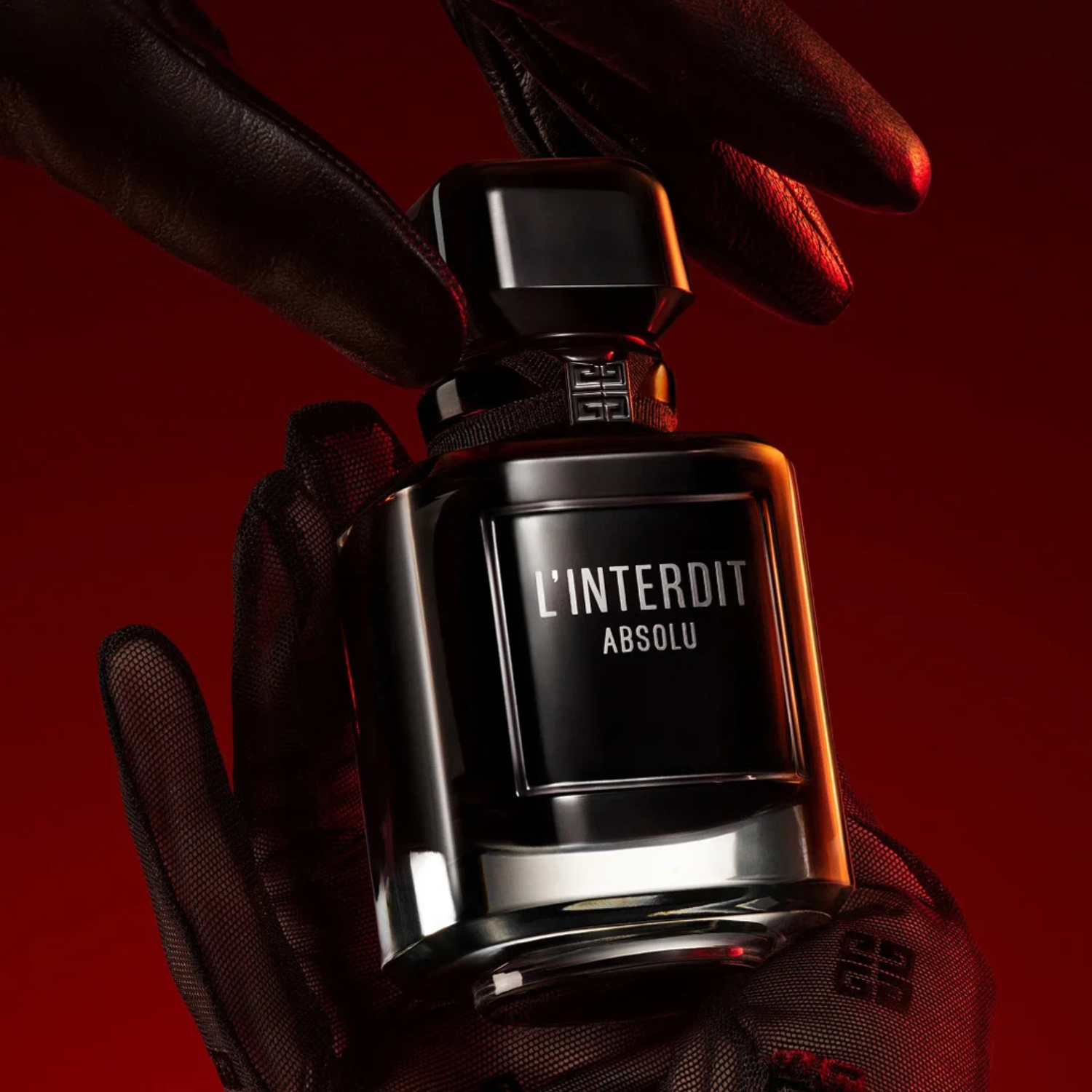 Perfume L’Interdit Absolu sendo segurado por luvas pretas, sobre fundo vermelho escuro, com iluminação suave que destaca o brilho do frasco.