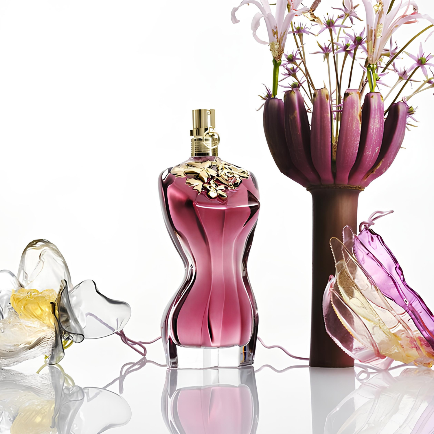 Perfume feminino La Belle Jean Paul Gaultier em destaque em um ambiente iluminado, cercado por flores exóticas em tons de rosa e roxo, e elementos translúcidos, transmitindo uma fragrância floral e sensual.