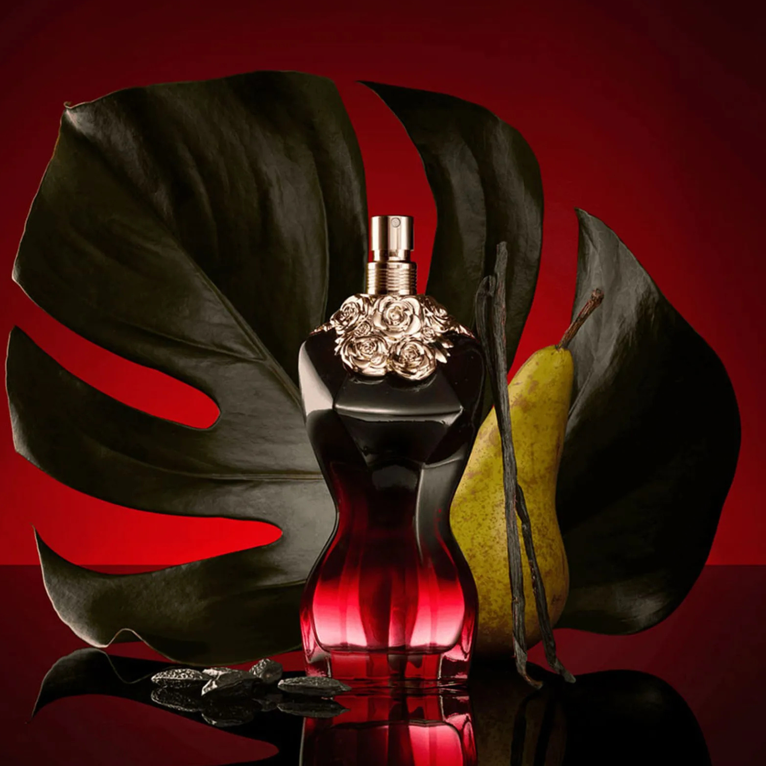 Perfume La Belle Le Parfum Jean Paul Gaultier em destaque em um cenário com uma grande folha de monstera escura, uma pera verde e favas de baunilha, sobre um fundo vermelho, reforçando as notas tropicais e amadeiradas da fragrância.