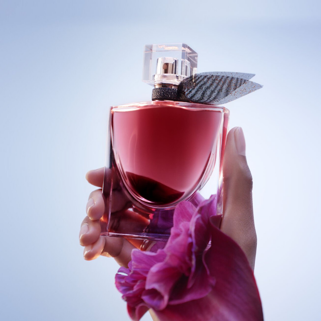 Mãos segurando o frasco de La Vie Est Belle L'Elixir Lancôme com uma grande pétala de flor rosa em primeiro plano e um fundo azul claro, destacando a delicadeza e a intensidade da fragrância.