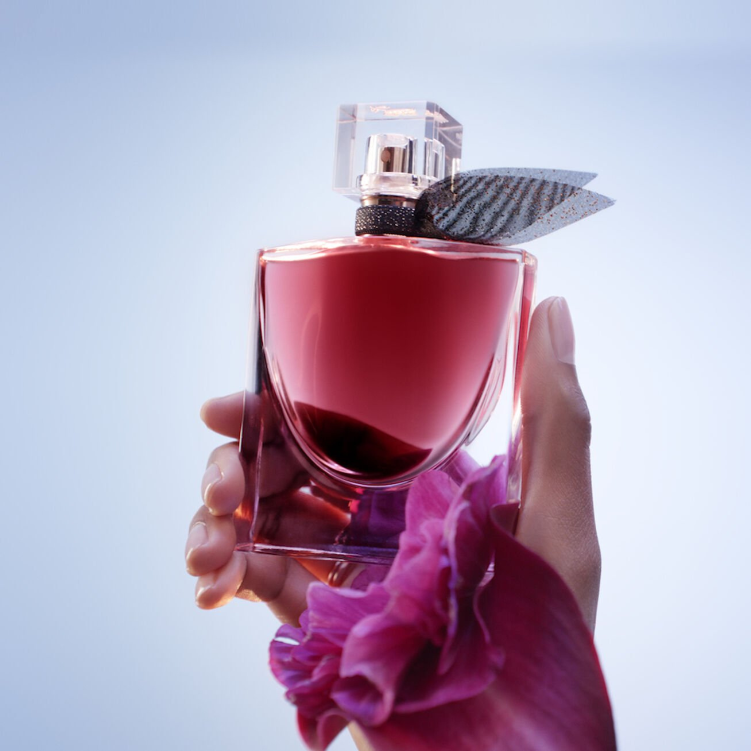 Mãos segurando o frasco de La Vie Est Belle L'Elixir Lancôme com uma grande pétala de flor rosa em primeiro plano e um fundo azul claro, destacando a delicadeza e a intensidade da fragrância.