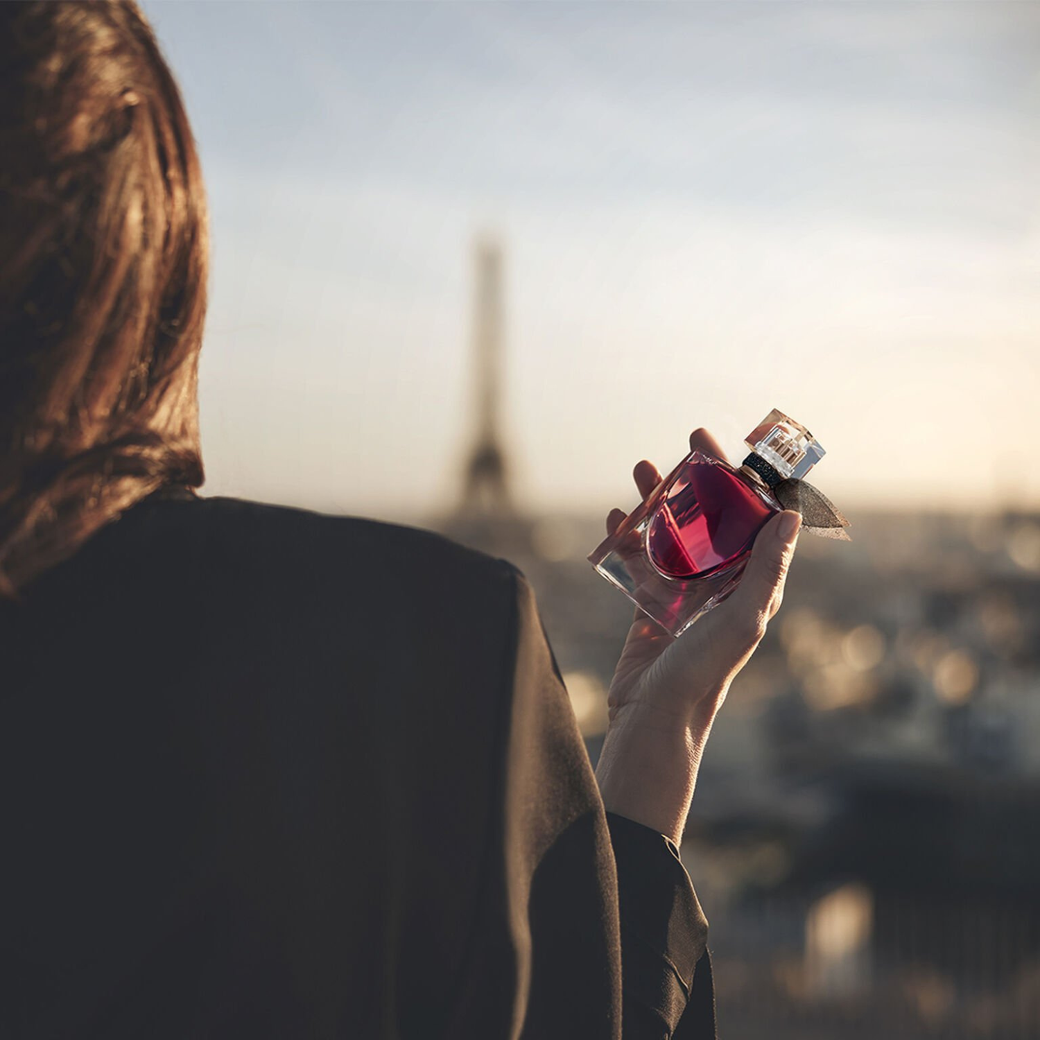 Visão de trás de uma mulher segurando delicadamente o frasco de La Vie Est Belle L'Elixir Lancôme com a Torre Eiffel ligeiramente desfocada ao fundo, transmitindo uma conexão com a icônica cidade e a beleza da vida.