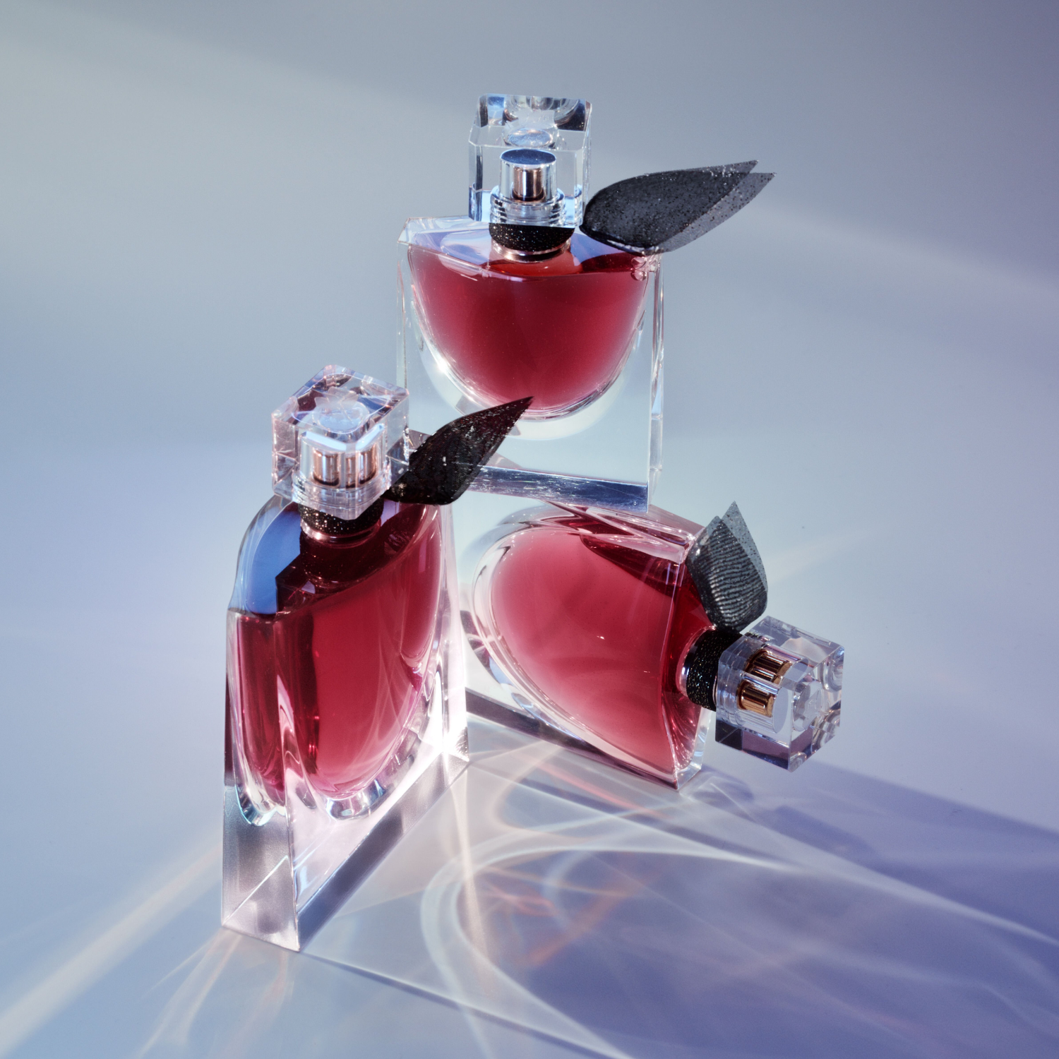 Três frascos do perfume La Vie Est Belle L'Elixir Lancôme dispostos sobre uma superfície clara com sombras suaves, realçando o design elegante e o tom rico da fragrância.