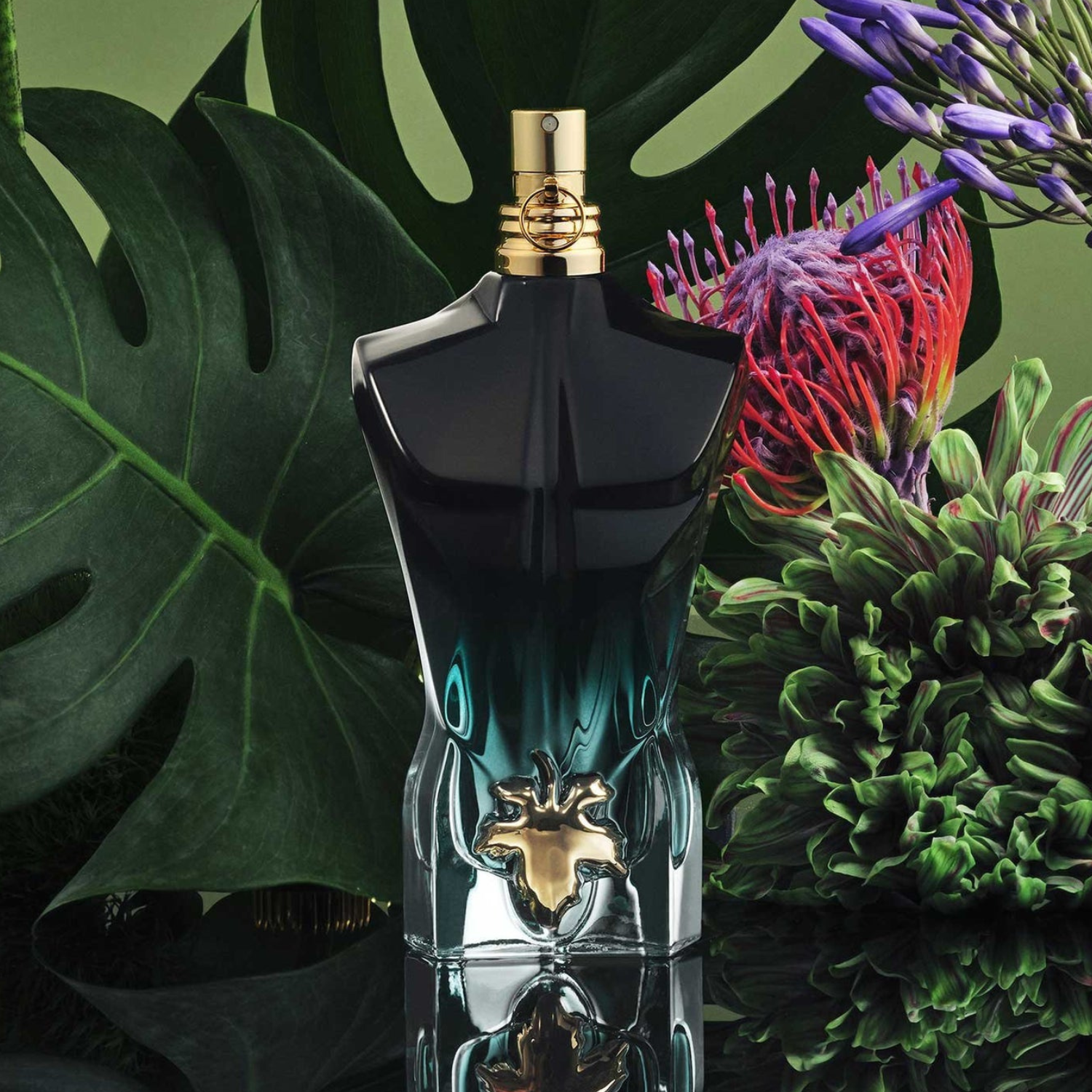 Perfume Le Beau Le Parfum Jean Paul Gaultier em destaque com uma grande folha verde ao fundo e flores tropicais coloridas, realçando a vitalidade e a exuberância da fragrância.