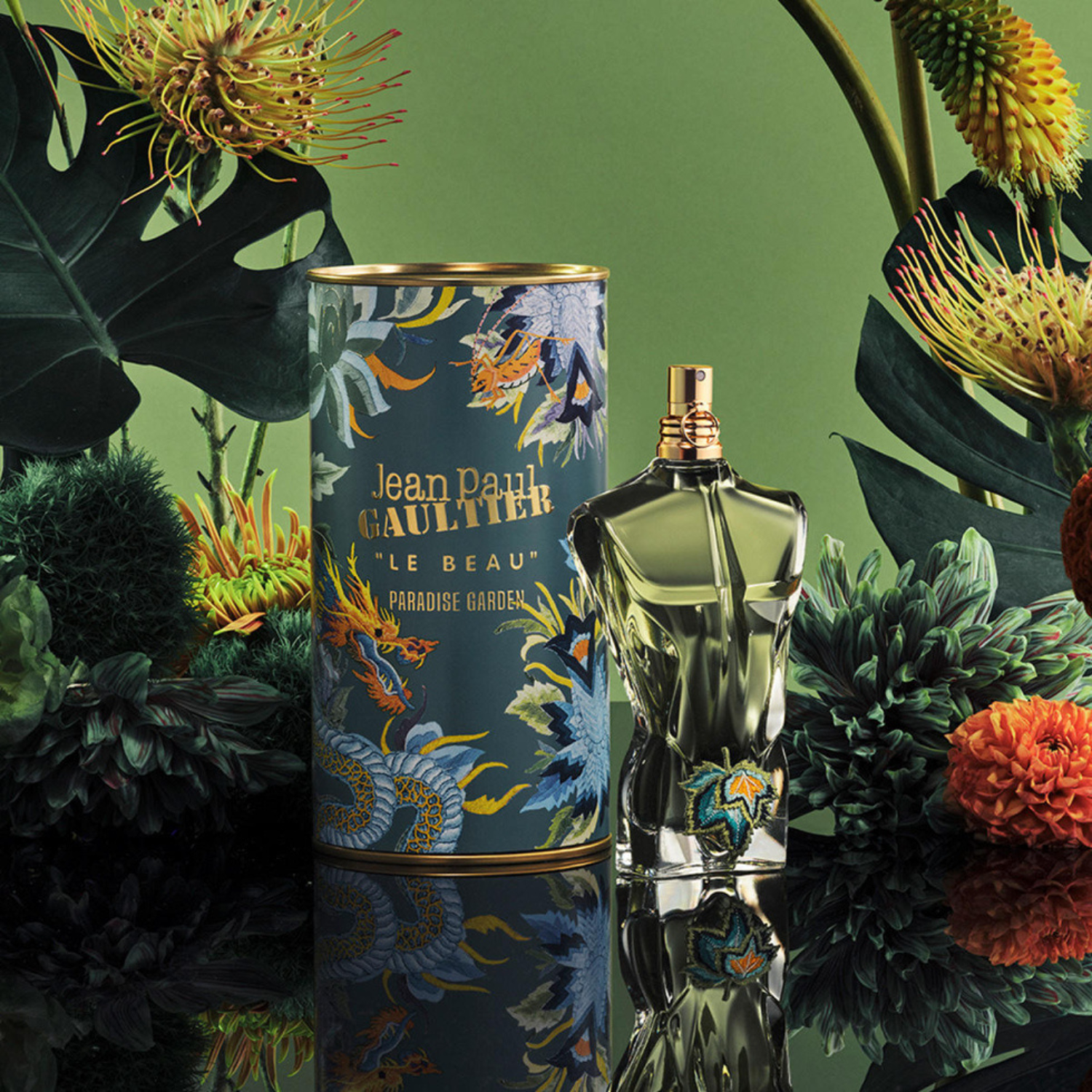 Frasco do perfume Le Beau Paradise Garden posicionado ao lado de sua embalagem cilíndrica decorada com ilustrações florais e dragões. Ambos estão inseridos em um cenário tropical com plantas exóticas e fundo verde.