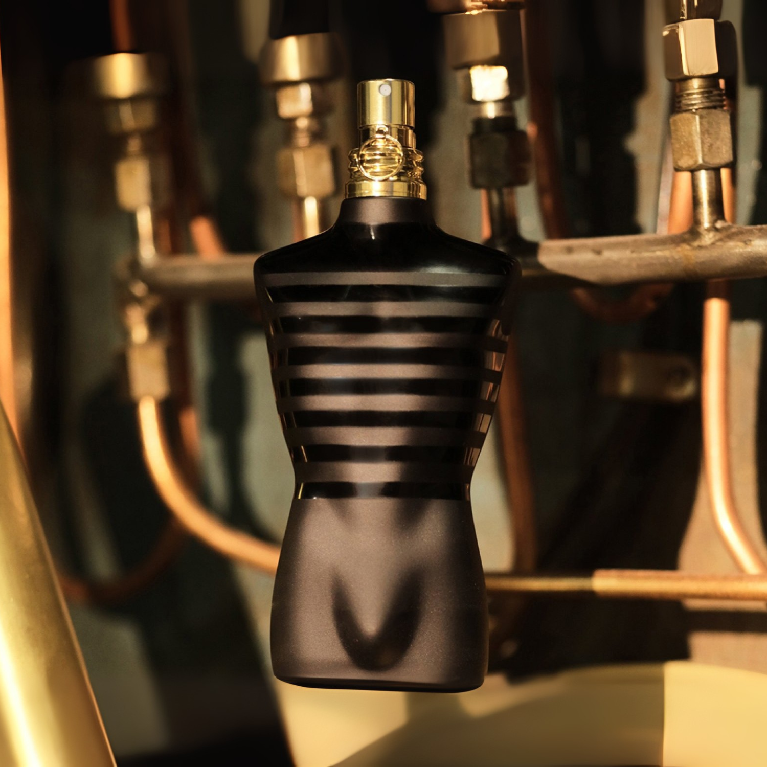 Perfume masculino Le Male Le Parfum Jean Paul Gaultier em destaque em um ambiente com tubulações e conexões metálicas em tons de bronze e prata ao fundo, criando uma atmosfera industrial e masculina.