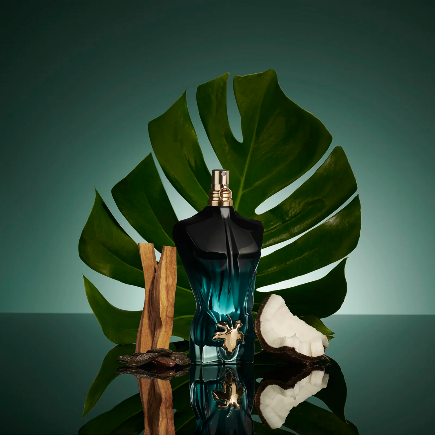 Perfume Le Beau Le Parfum Jean Paul Gaultier em destaque sobre uma superfície reflexiva, com uma folha de monstera, pedaços de madeira e coco ao redor, reforçando as notas tropicais e amadeiradas da fragrância.