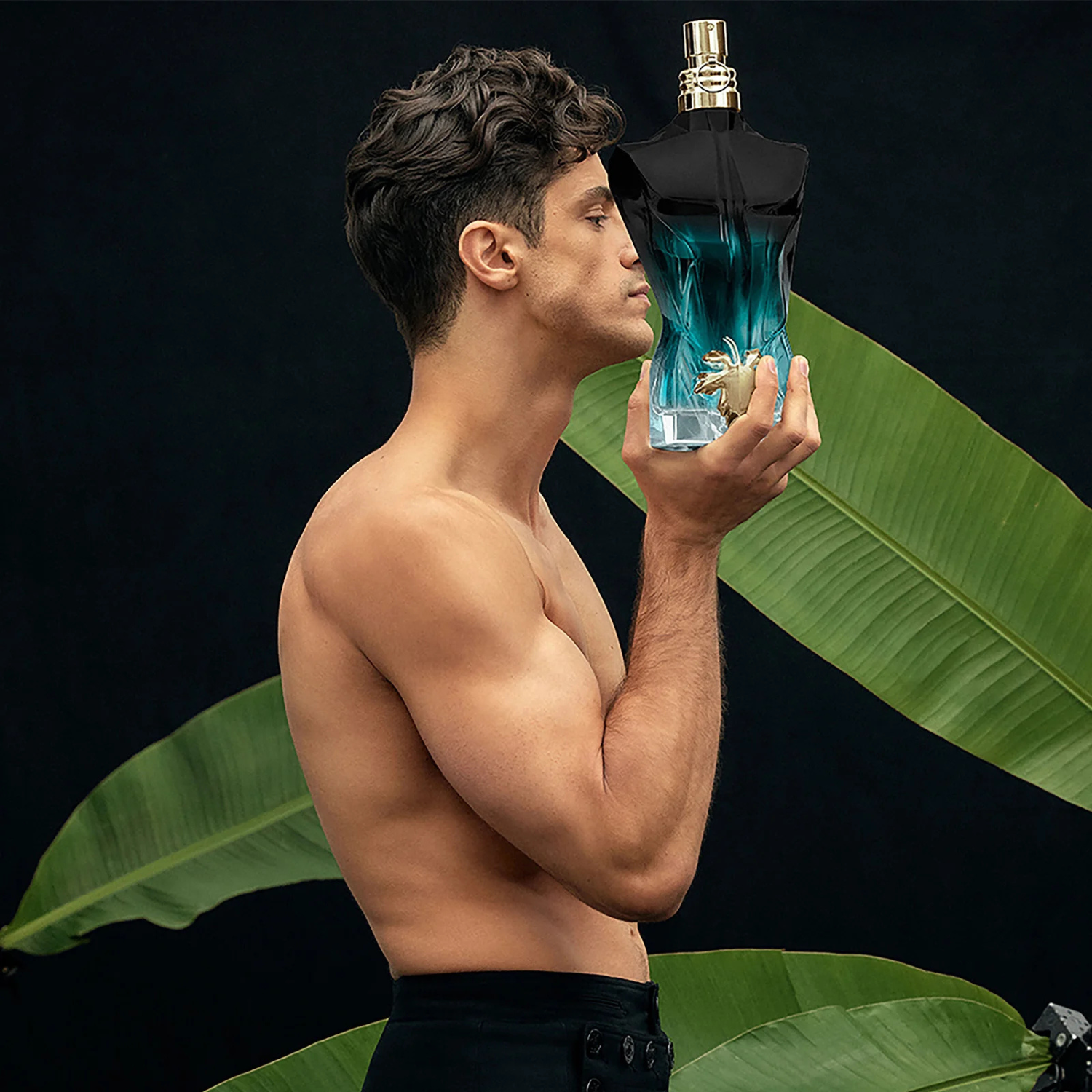 Modelo masculino segurando o perfume Le Beau Le Parfum Jean Paul Gaultier próximo ao rosto, com grandes folhas verdes ao fundo, sugerindo a conexão da fragrância com a natureza e a sensualidade.
