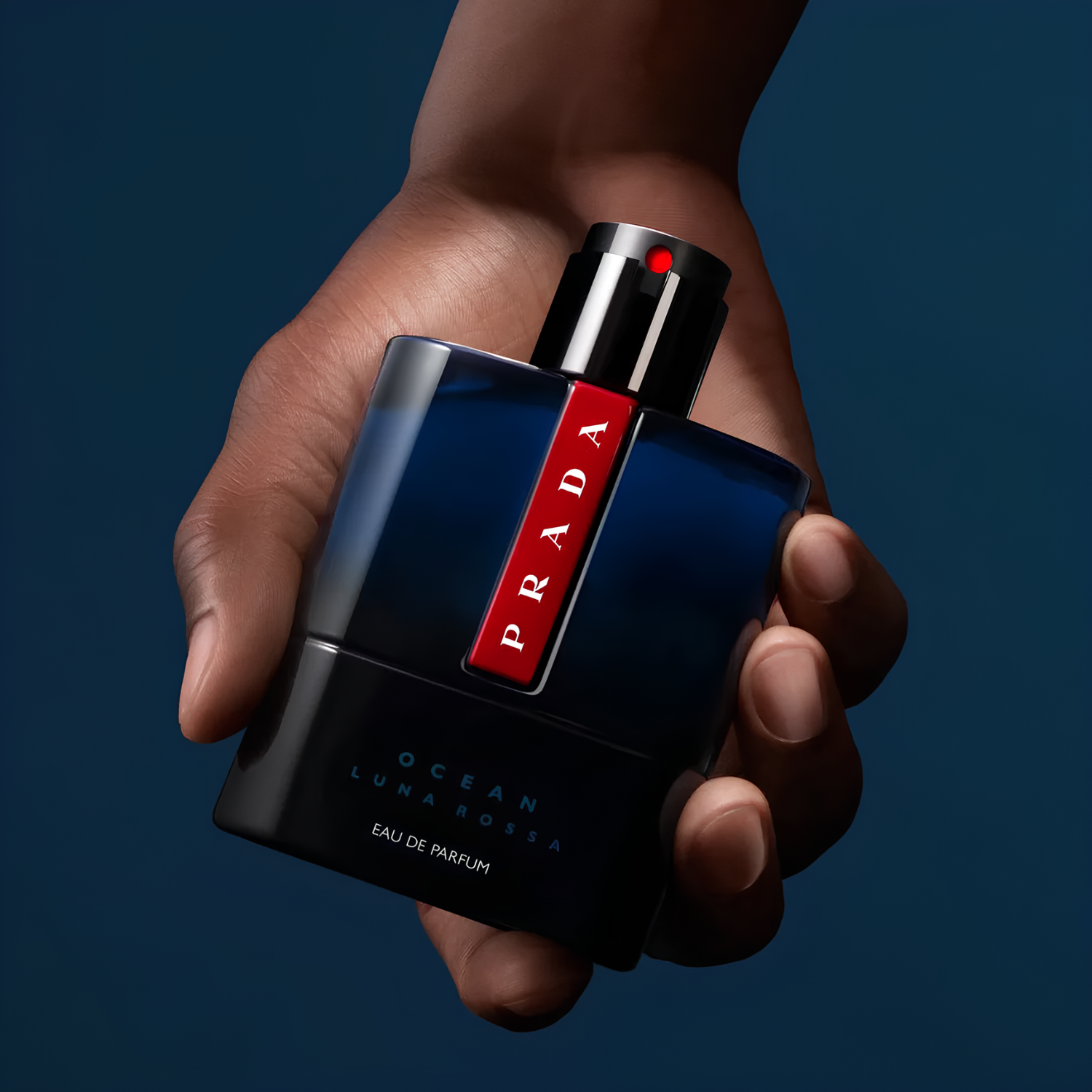 Perfume Prada Luna Rossa Ocean sendo segurado por uma mão masculina contra fundo azul escuro, transmitindo força e elegância.