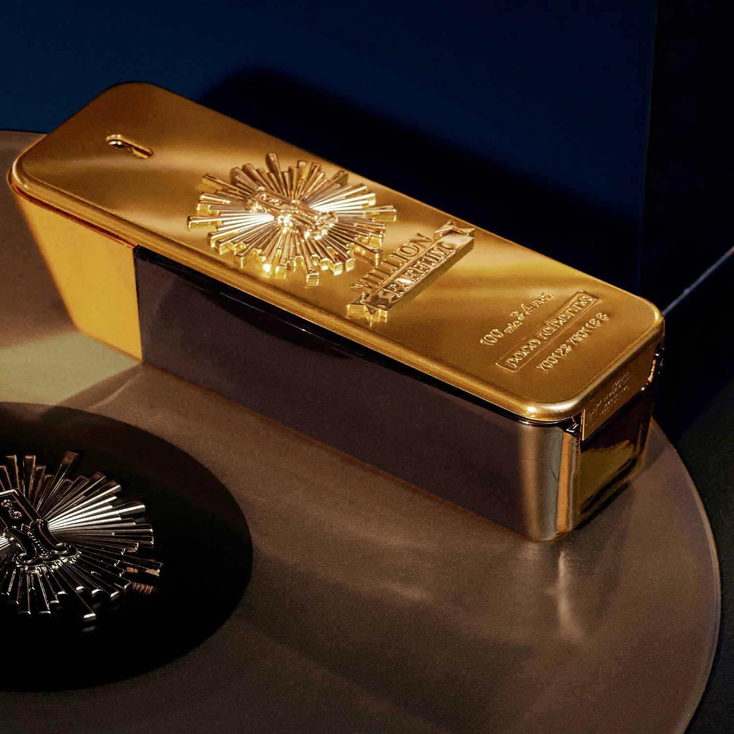 Perfume importado masculino One Million Parfum Rabanne posicionado sobre uma superfície escura e reflexiva, com outro frasco similar desfocado ao lado, enfatizando o brilho do ouro em contraste com o fundo, disponível na Velyre.