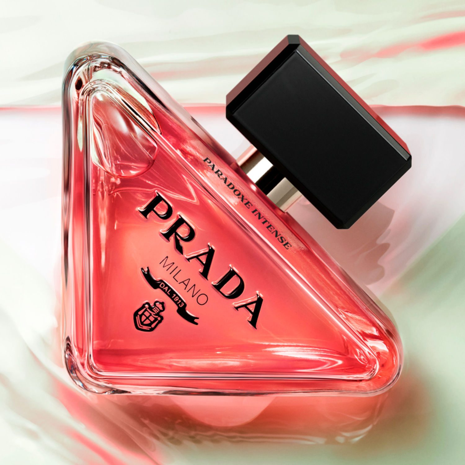 Perfume Paradoxe Intense Prada posicionado em um fundo com textura em tons de rosa e branco, com a tampa preta ao lado, destacando o design triangular do frasco e a cor vibrante da fragrância.