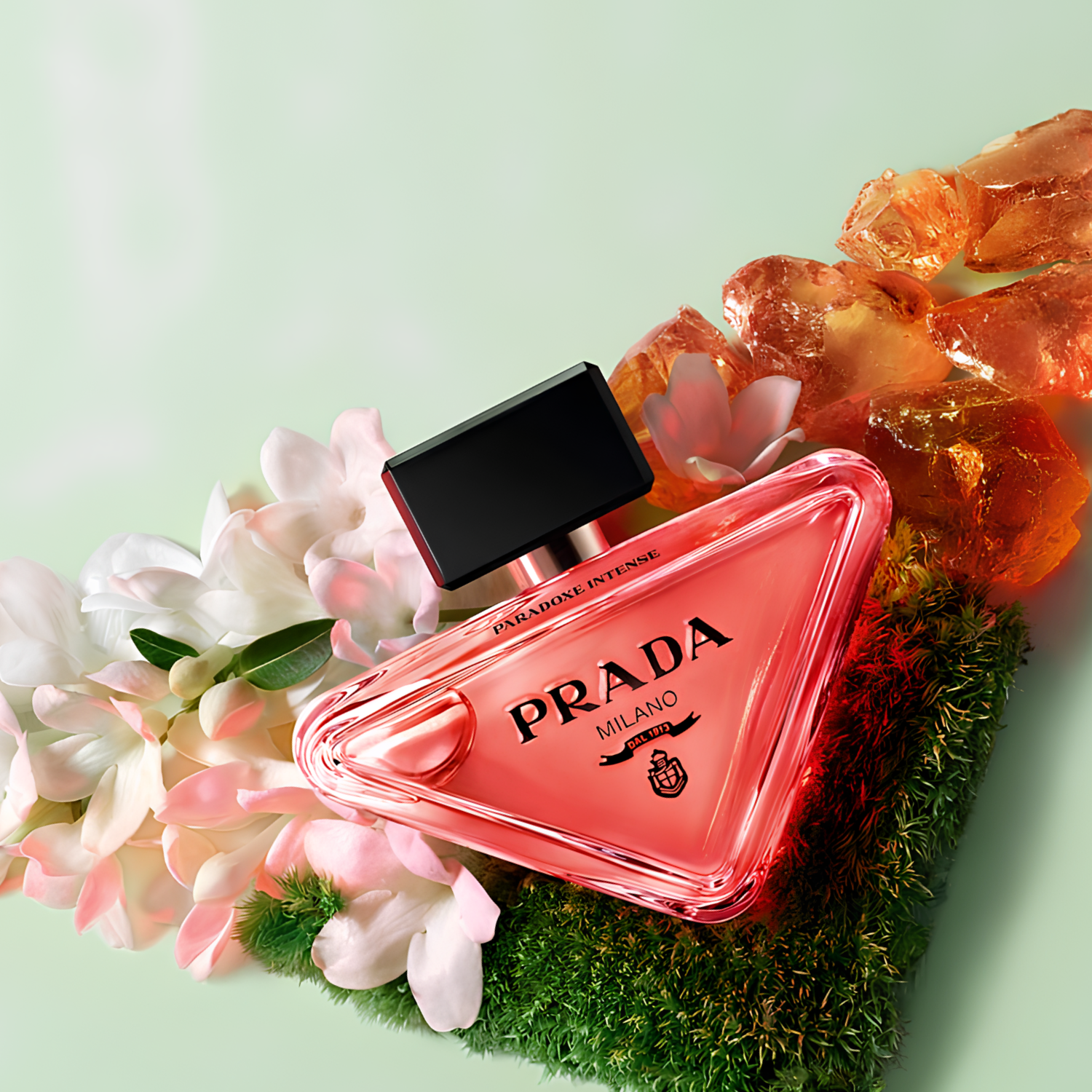 Perfume feminino Paradoxe Intense Prada em destaque em uma composição com delicadas flores brancas, elementos âmbar cristalizados e musgo, sugerindo uma fragrância floral com toques quentes e luminosos.