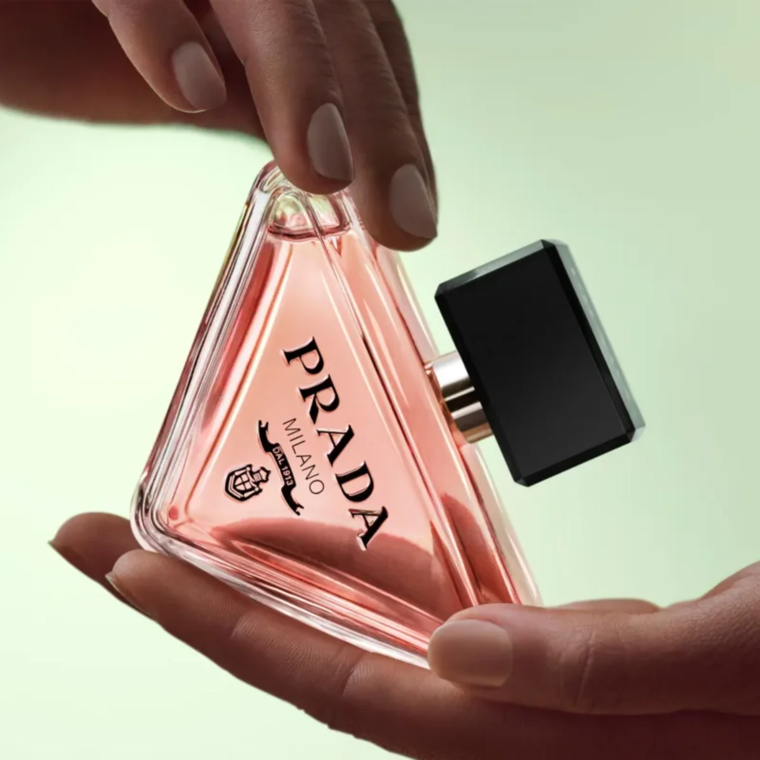 Mãos femininas delicadamente segurando o frasco triangular do perfume Paradoxe Prada, com unhas em tons neutros, sobre um fundo verde suave, transmitindo uma sensação de cuidado e apreço pelo perfume.