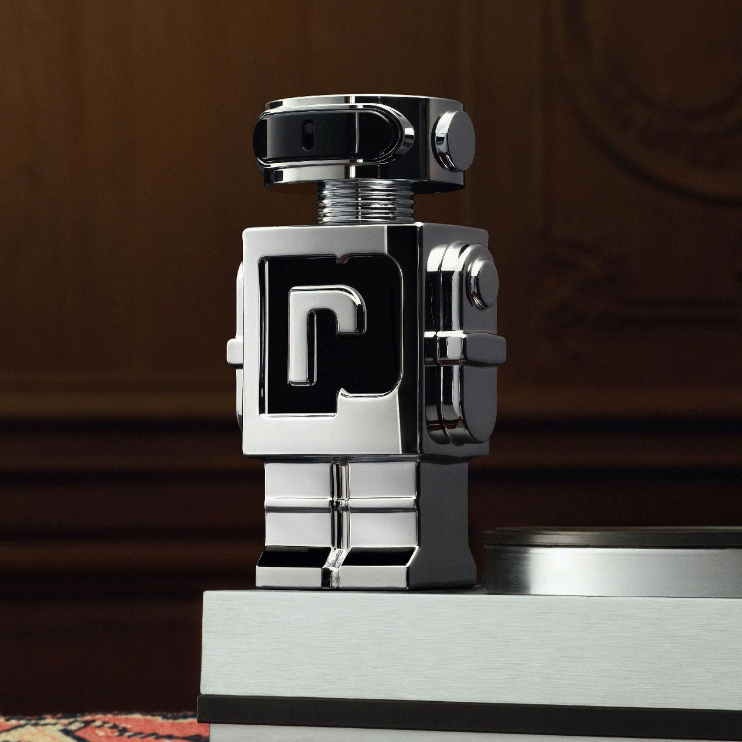 Perfume importado masculino Phantom Rabanne posicionado sobre um livro de capa clara, com fundo escuro, realçando o brilho metálico do frasco robô e transmitindo uma sensação de inovação e futurismo, disponível na Velyre.