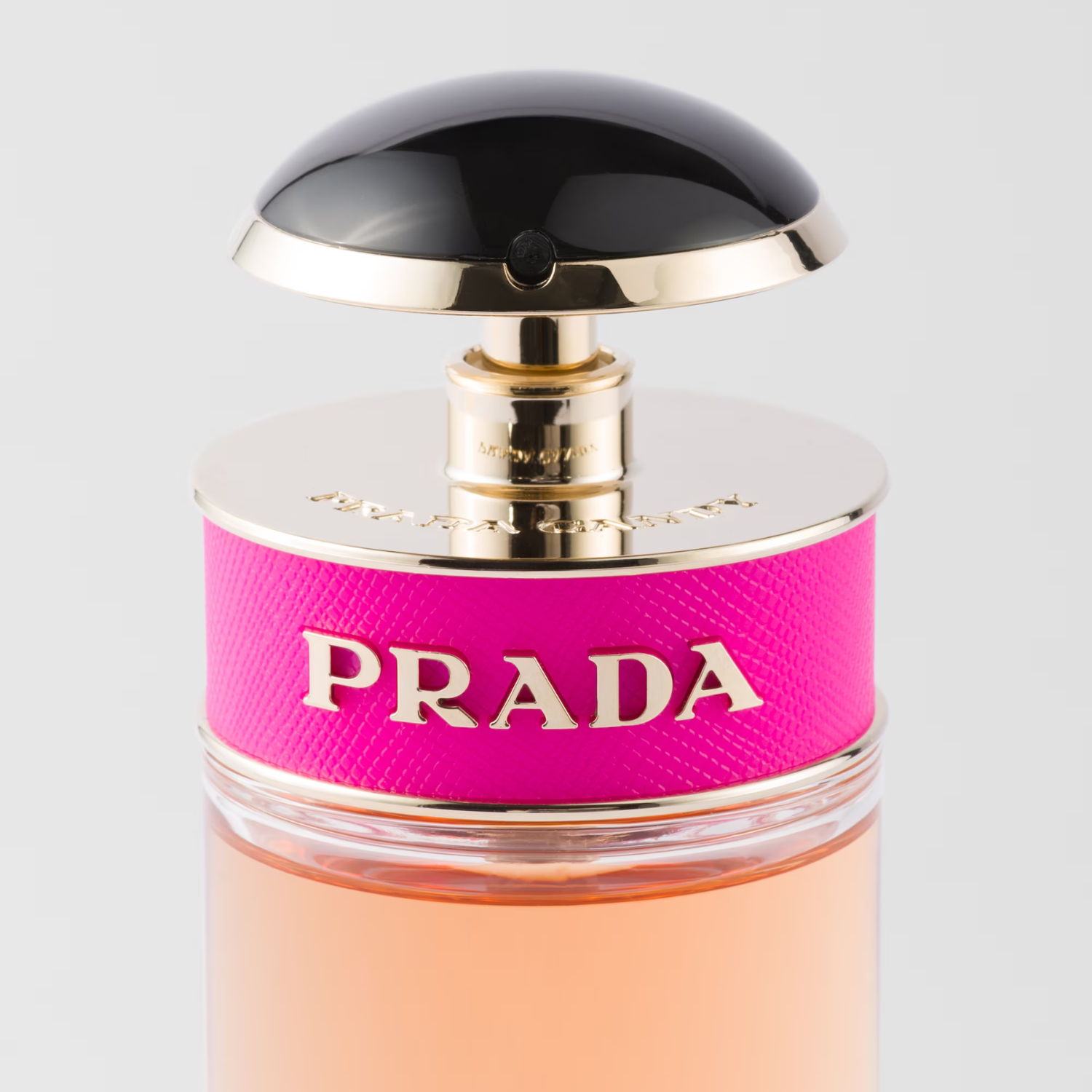 Close-up da parte superior do frasco do perfume Prada Candy Prada, destacando a faixa de couro rosa choque com a inscrição "PRADA" em dourado e a tampa em formato de cúpula preta e dourada.