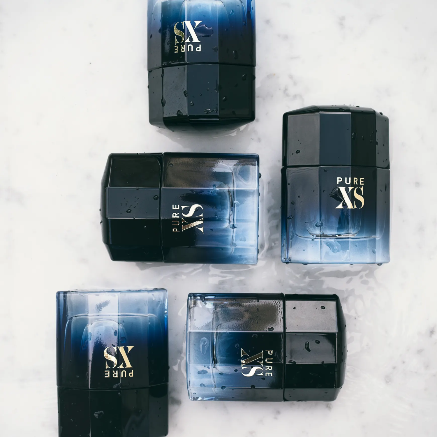 Vista aérea de múltiplos frascos do perfume Pure XS Rabanne dispostos sobre uma superfície clara, alguns com gotas de água, realçando o design uniforme e a sensação sensual da fragrância.