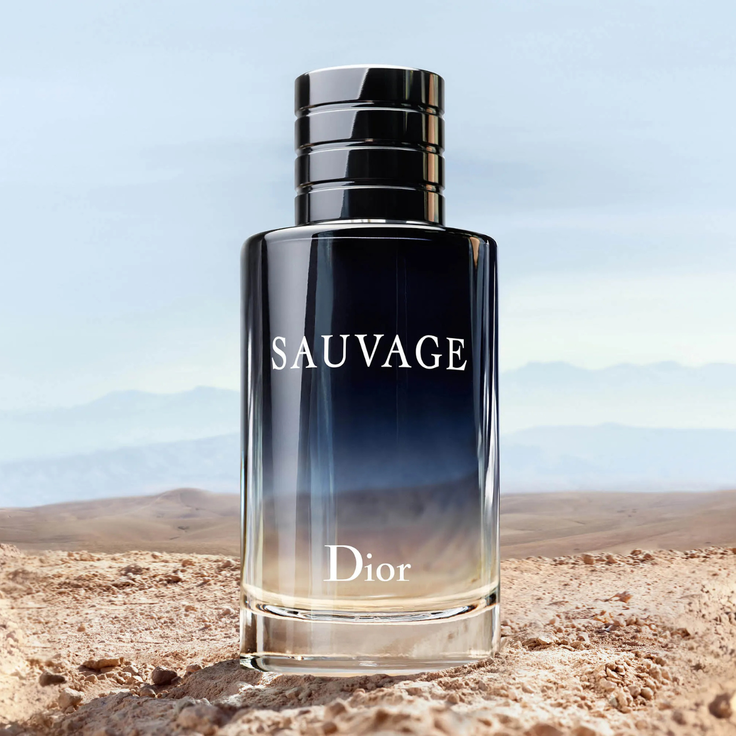 Perfume masculino Sauvage Dior em destaque sobre uma superfície arenosa com montanhas ao fundo e céu azul claro, evocando uma sensação de liberdade e natureza selvagem.
